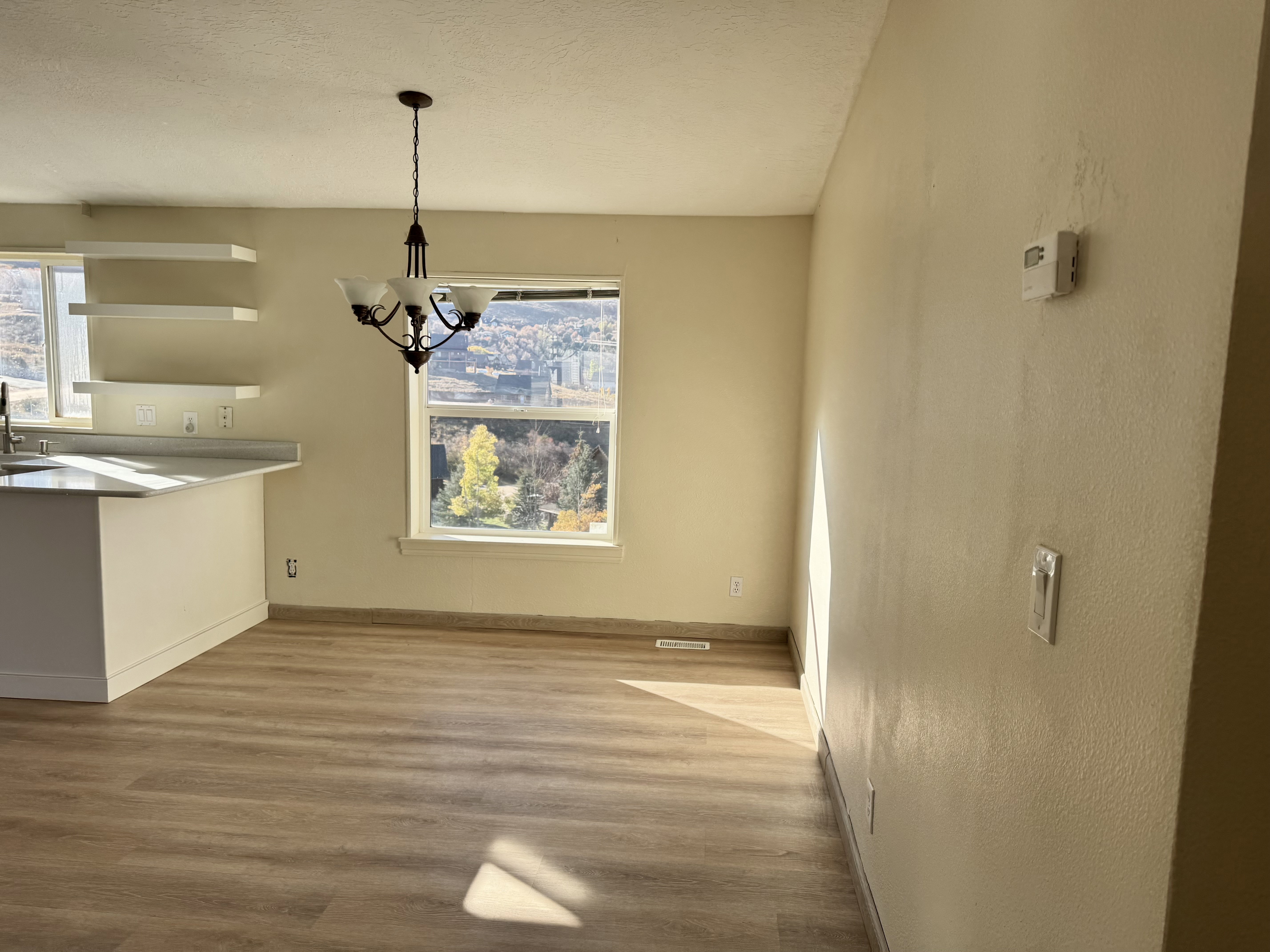 Open-concept area featuring wood-finish flooring, a contemporary chandelier, a kitchen island with a light-colored countertop, and a window framing a distant landscape