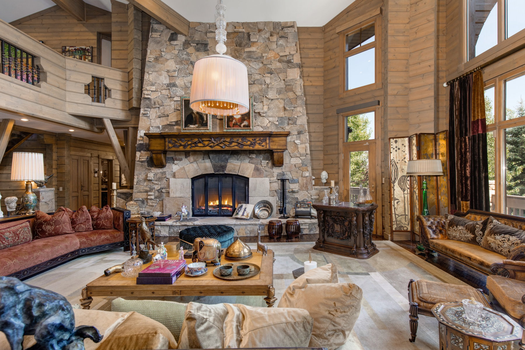 Spacious living area featuring a stone fireplace with a carved wood mantel, high wood-paneled ceilings, a second-story interior balcony, and large windows with exterior foliage