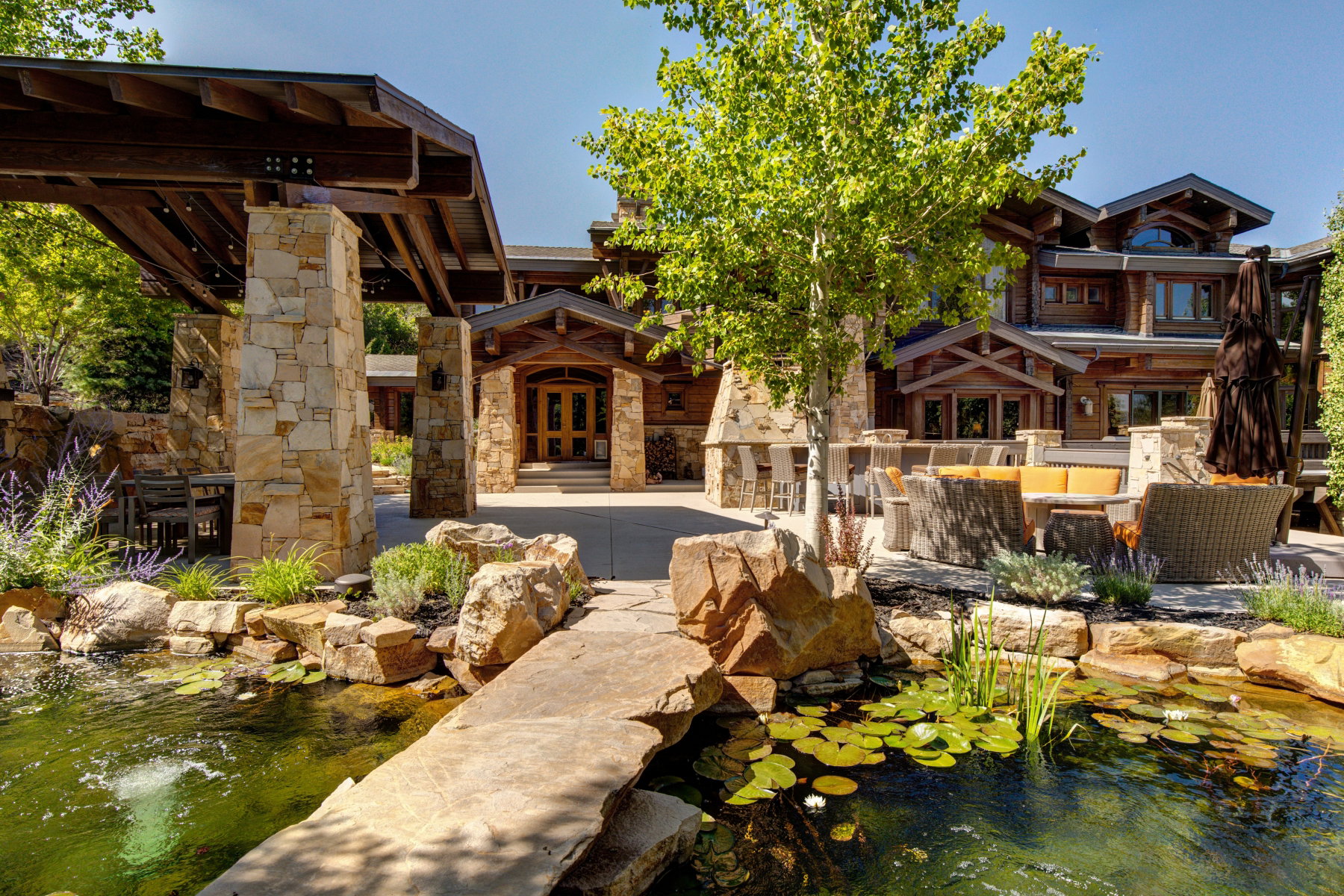 Expansive custom residence featuring a stone-clad exterior with wood accents, a stone patio with a built-in fireplace, a covered outdoor dining area, and a tranquil pond with a stone pathway