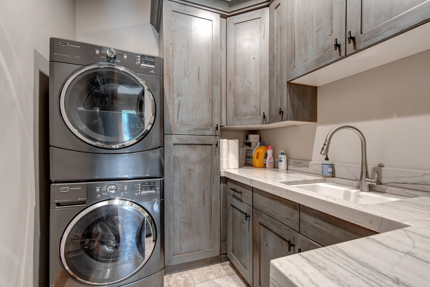 Laundry area with stacked appliances, light wood-finish cabinetry, and stone countertops featuring an undermount sink and gooseneck faucet