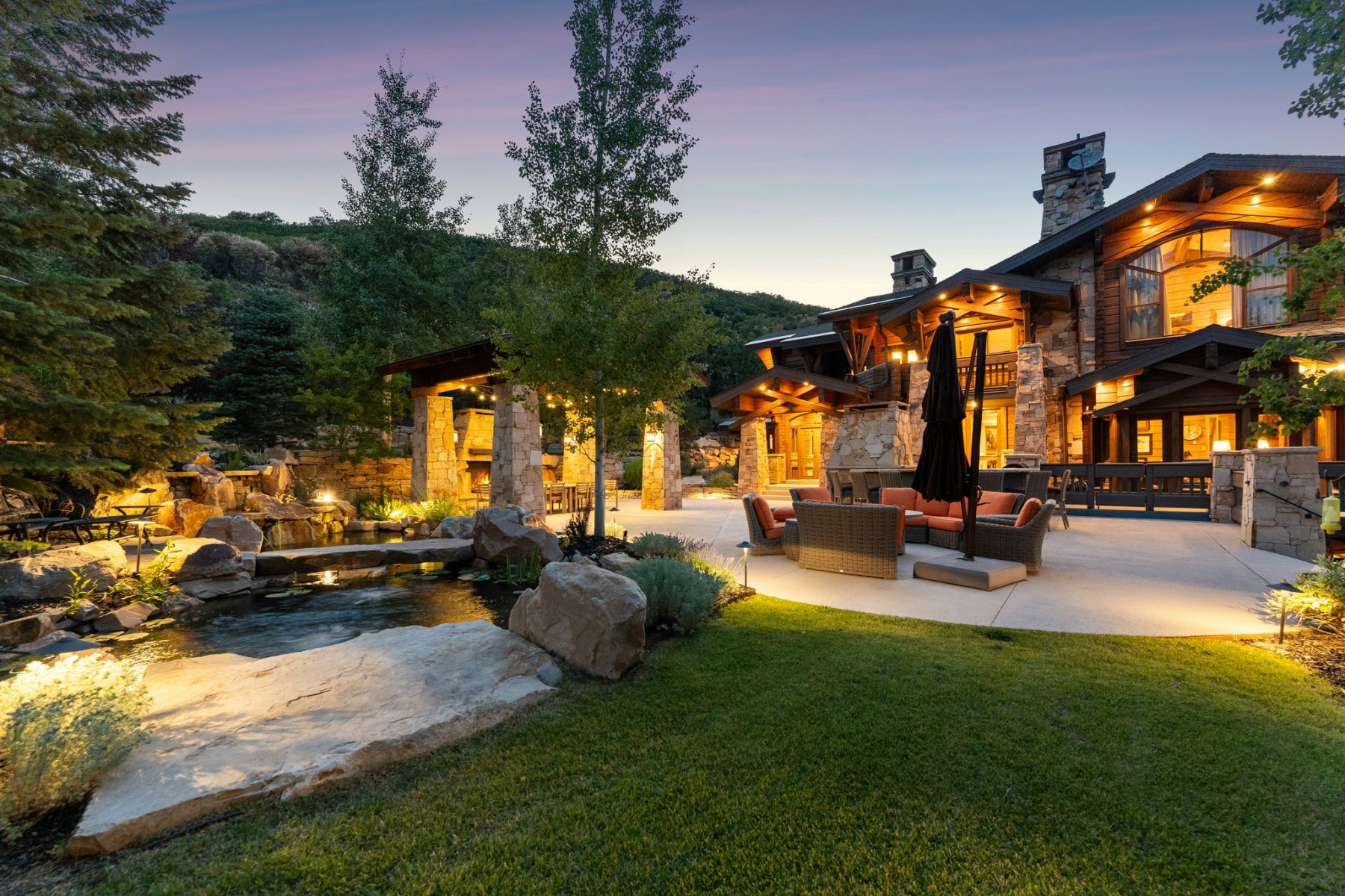 Stone and timber exterior featuring a spacious patio with integrated lighting, an outdoor kitchen with a stone fireplace, and a naturalistic water feature