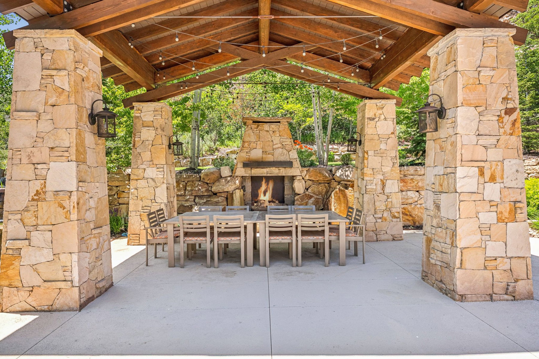 Expansive outdoor patio featuring a stone fireplace, wood-beamed ceiling, integrated lighting, stone support pillars, and concrete flooring