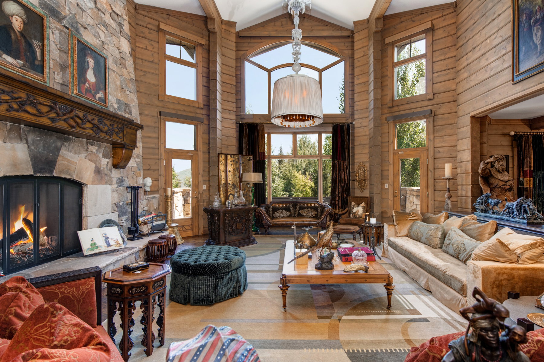 Grand living space featuring a two-story stone fireplace with an ornate wood mantel, natural wood paneling, floor-to-ceiling windows, soaring vaulted ceilings with exposed beams, and a large central chandelier