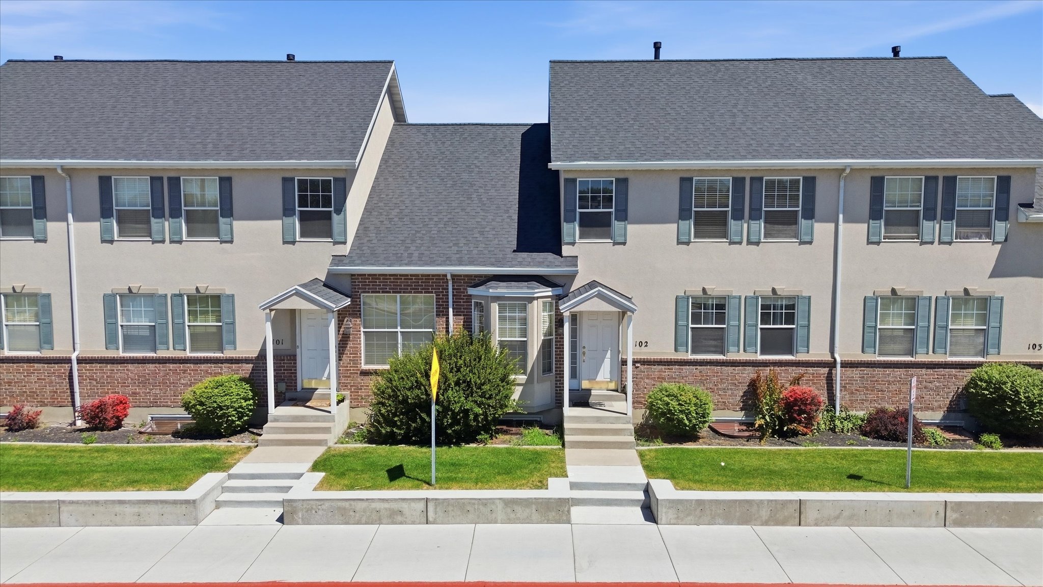 Townhouse exterior featuring a gray shingled roof, light-colored stucco siding, and a brick facade