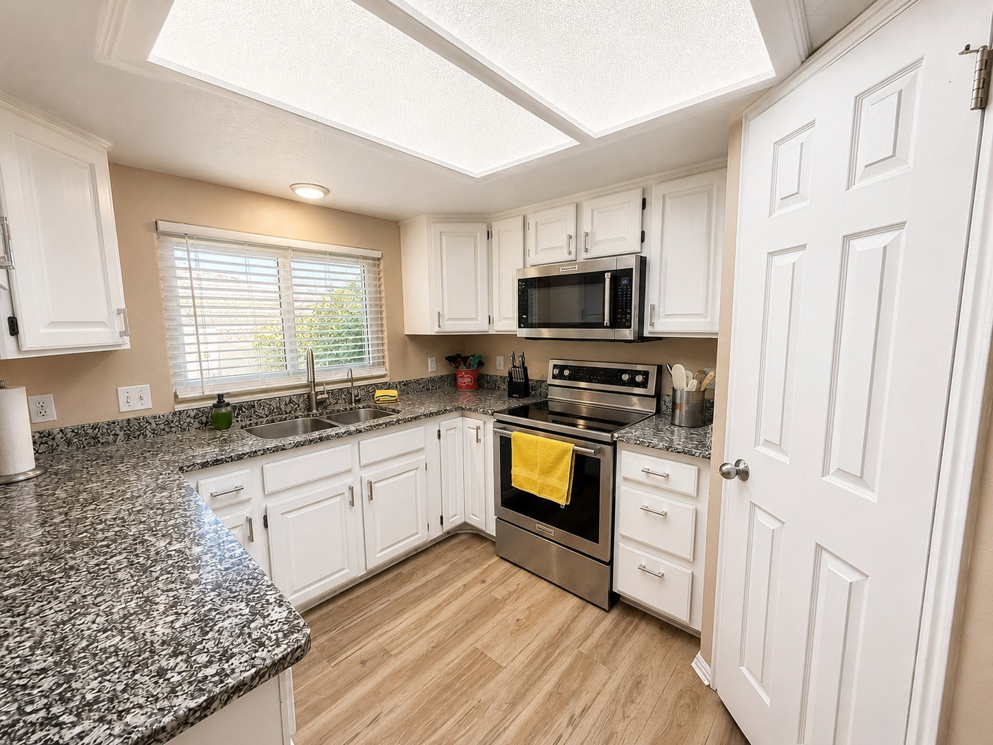 Kitchen featuring white cabinetry, speckled countertops, stainless steel appliances, wood-finish flooring, and a double basin sink beneath a window