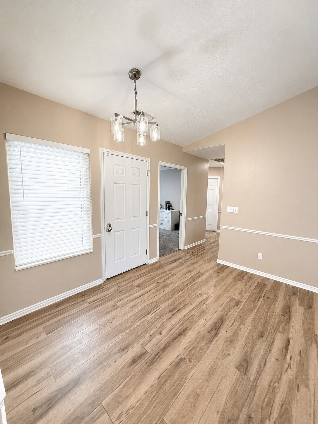 Spacious room featuring wood-finish flooring, a contemporary chandelier, a white interior door, and a large window with blinds