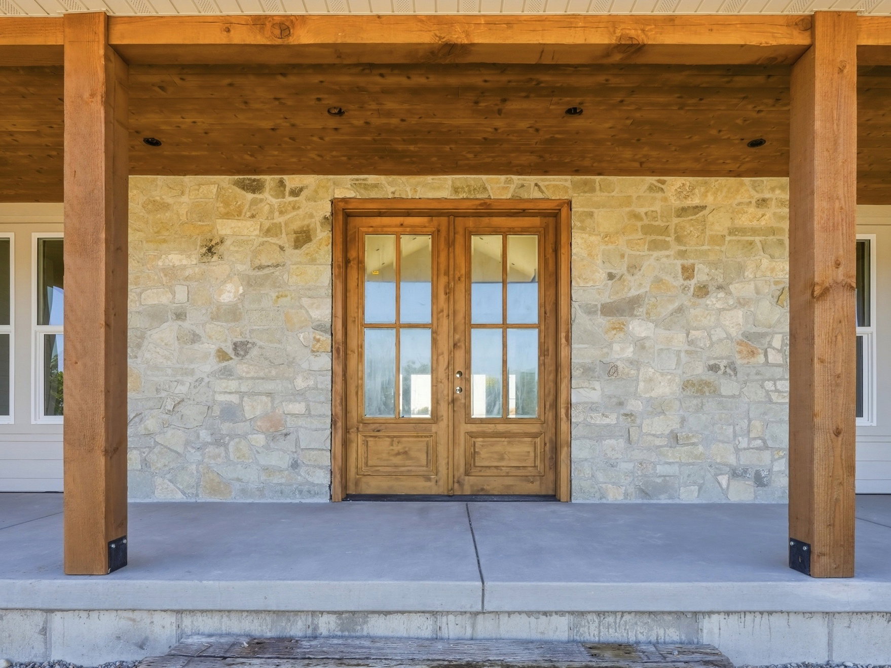 Stone facade with a covered concrete porch, featuring exposed wood beams and posts, and a double wood-paneled entry door with glass inserts