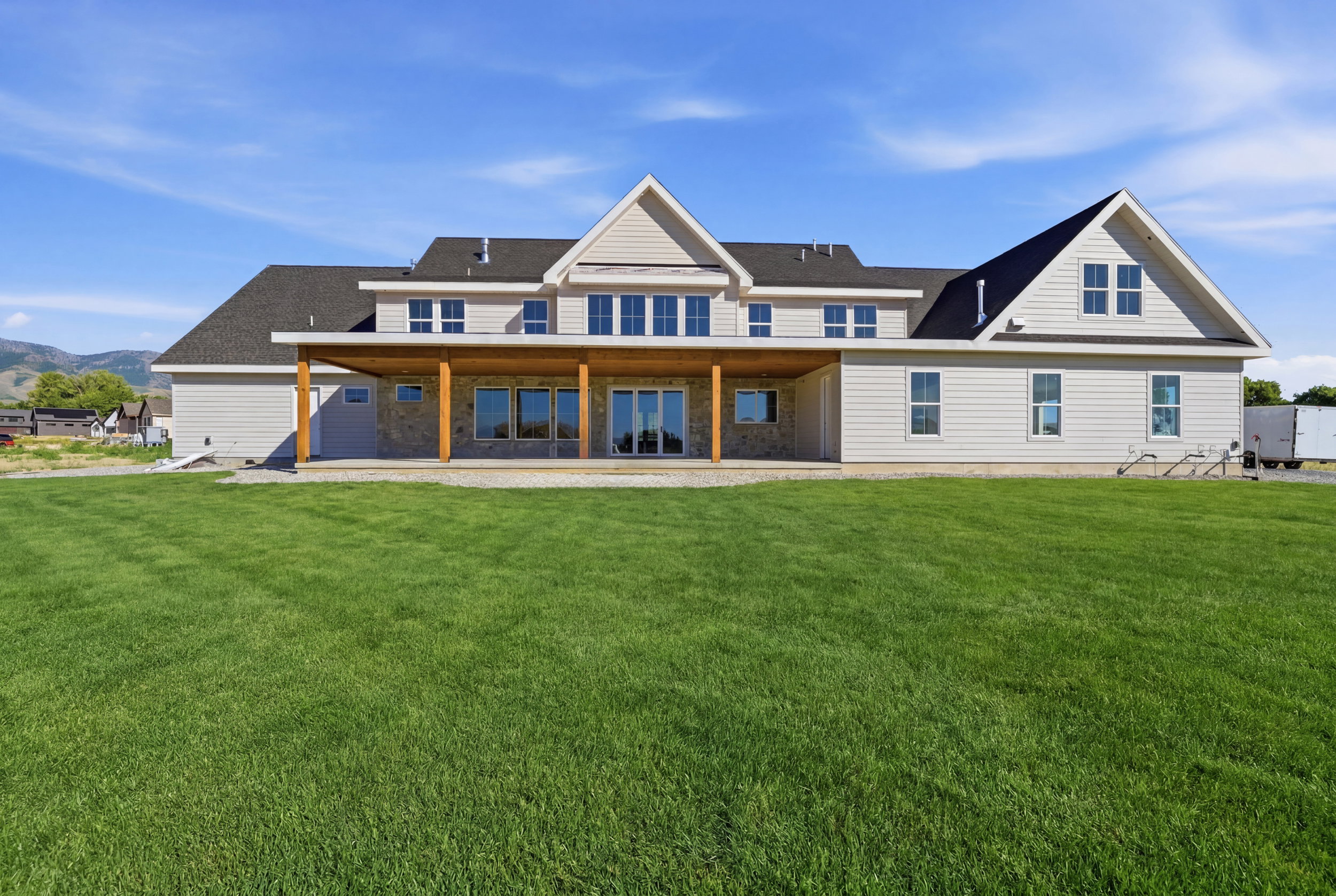 Expansive rear exterior featuring a covered patio with exposed wood beams, multiple large windows, a stone accent wall, and a broad green lawn