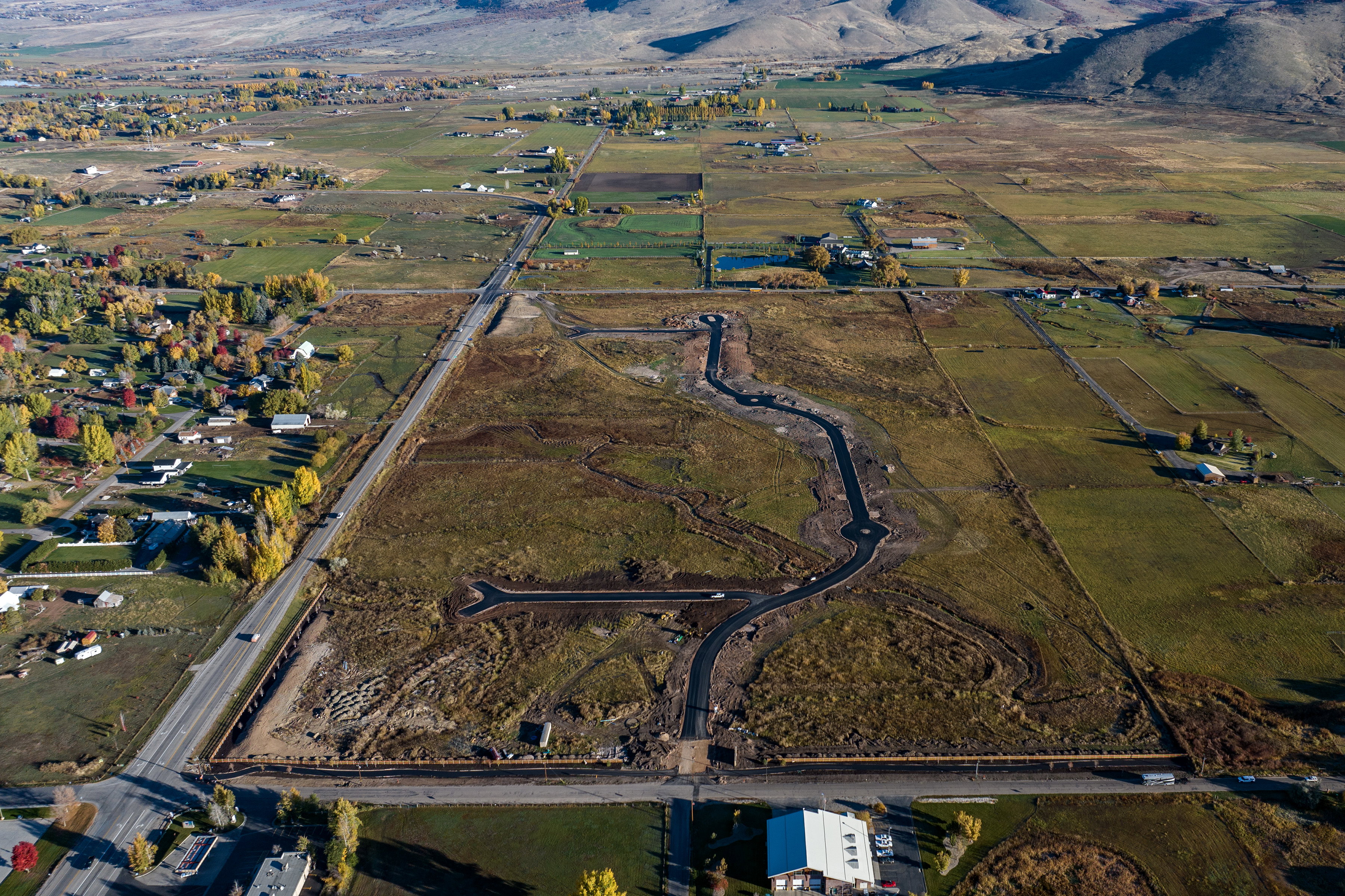 Expansive undeveloped land featuring newly constructed roadways and cul-de-sacs, surrounded by mature trees, open fields, and distant mountains