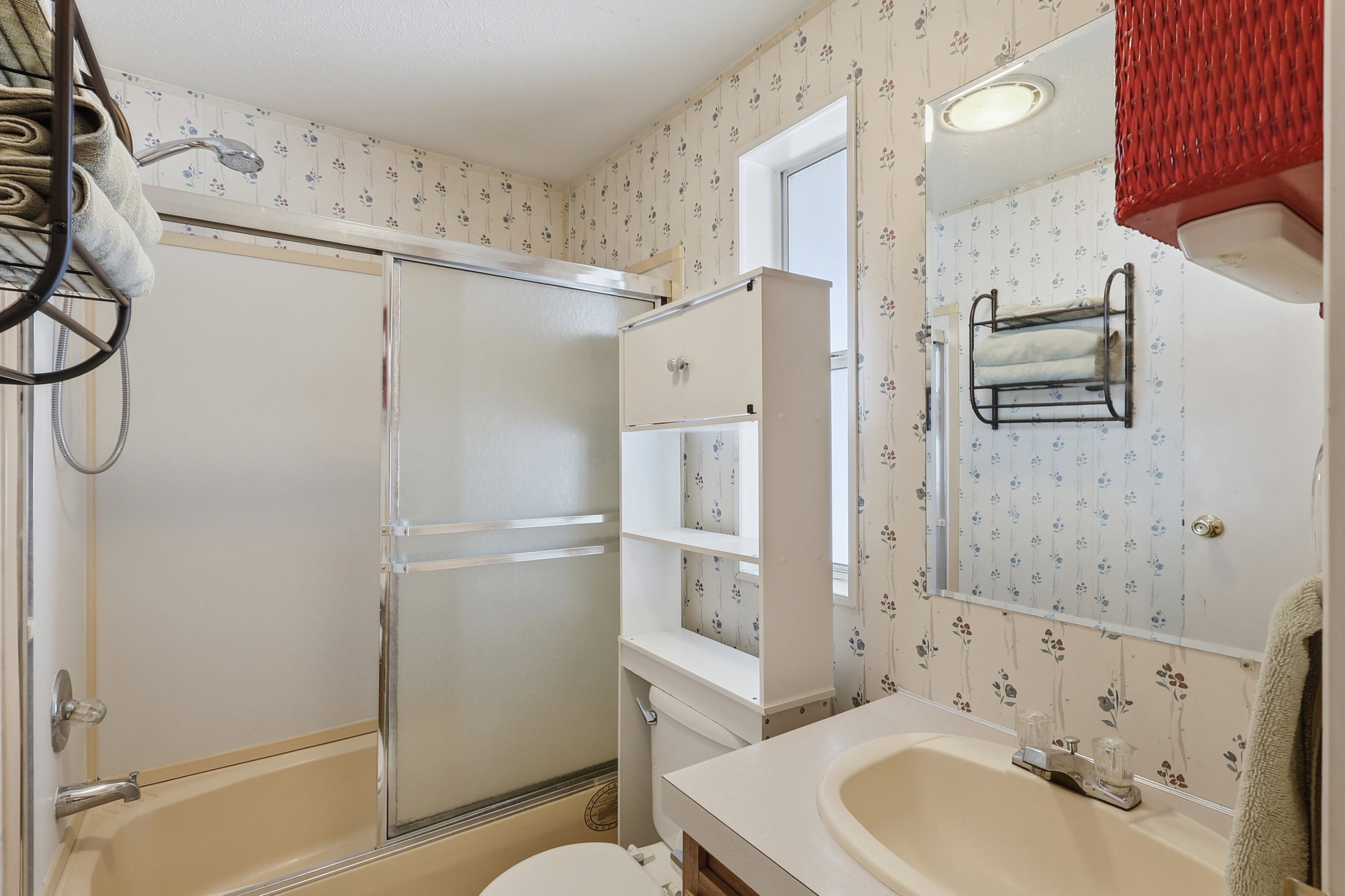 Bathroom featuring a bathtub with a sliding glass enclosure, a vanity with an integrated sink, a built-in wall cabinet with open shelving, a wall-mounted mirror, and a window for natural light