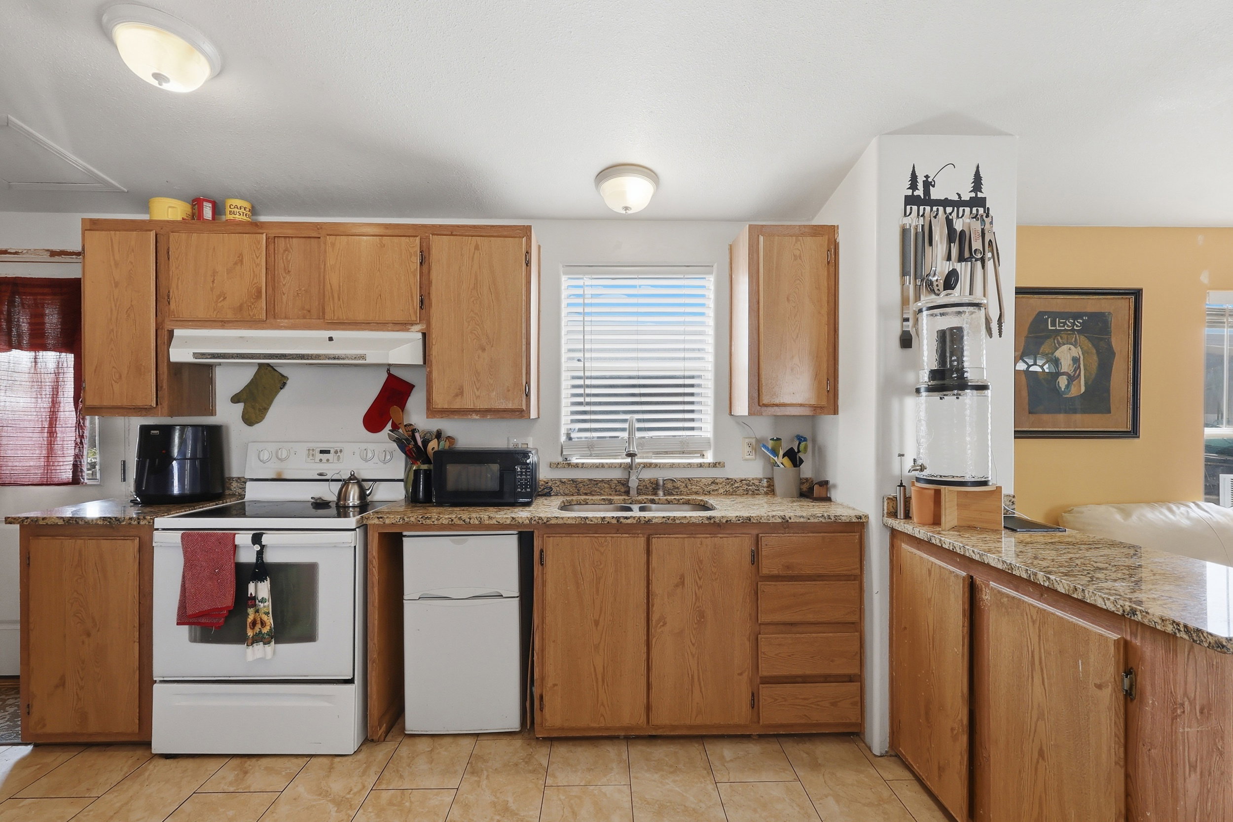 Kitchen featuring wood-finish cabinetry, light-toned granite-style countertops, white electric range with overhead vent, stainless steel sink with window above, and tiled flooring
