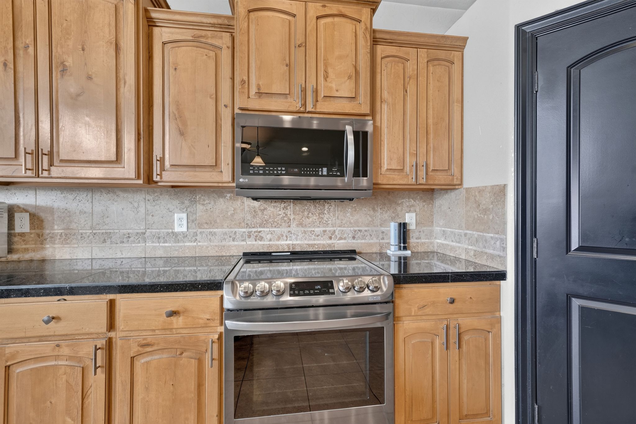 Kitchen featuring light wood cabinetry, stainless steel oven with an electric cooktop, built-in microwave, dark countertops, and a neutral tile backsplash