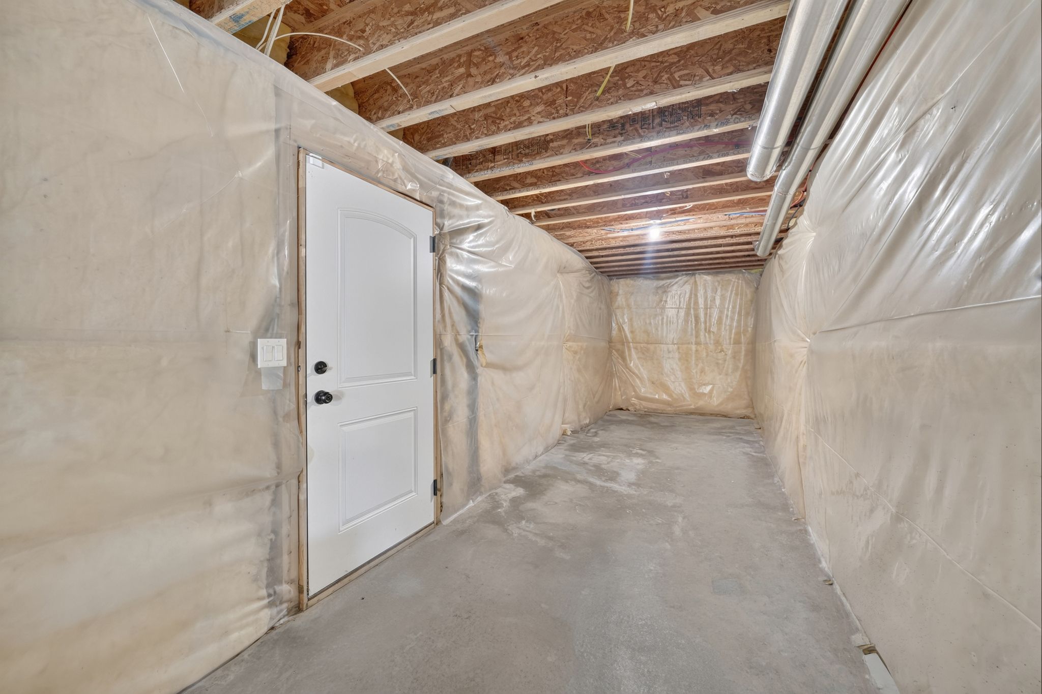Unfinished basement space featuring a white paneled door with black hardware, exposed ceiling joists, concrete flooring, and translucent wall coverings