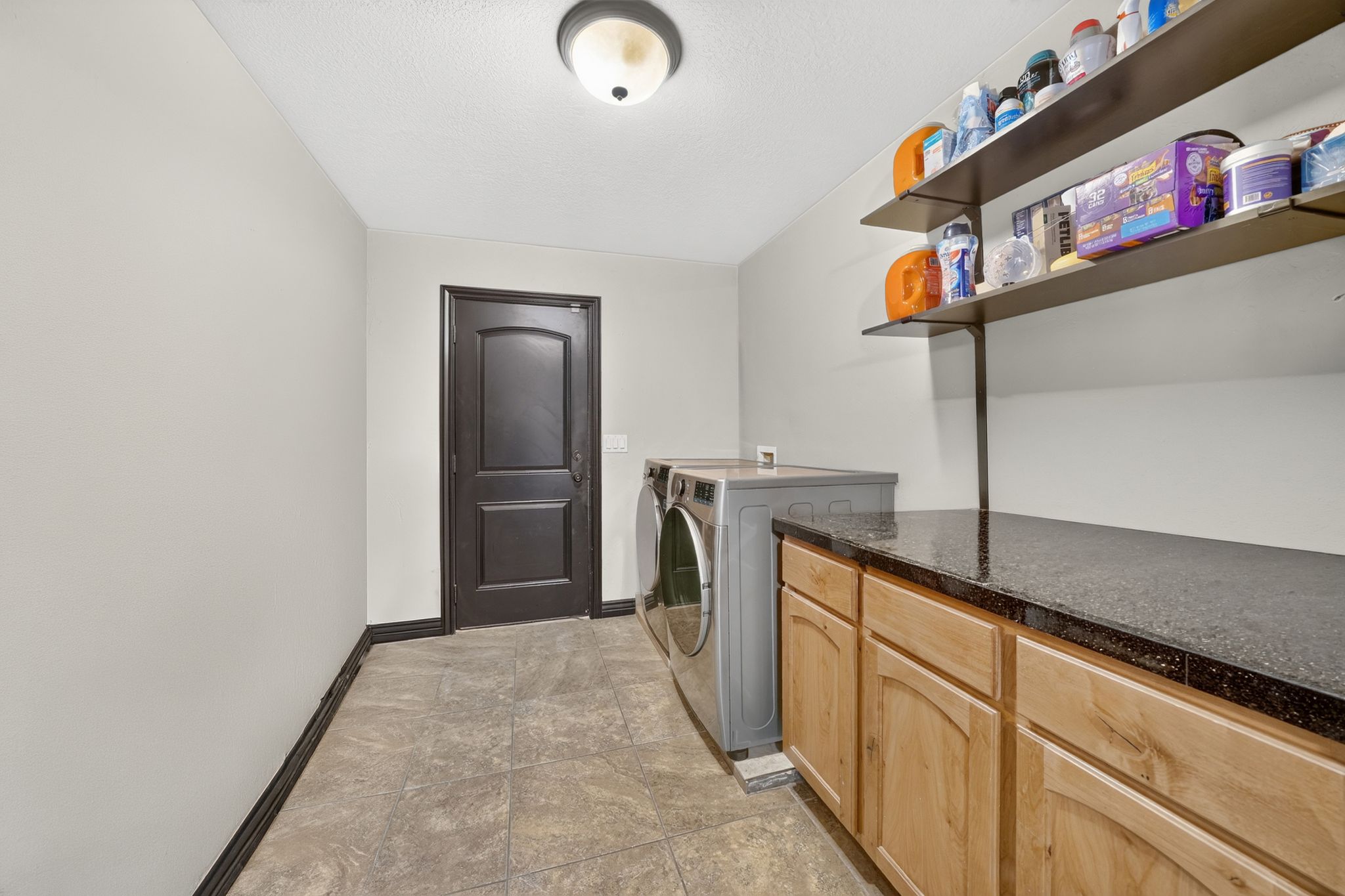 Laundry room featuring stone-finish tile flooring, a dark paneled door, light wood cabinetry, a dark speckled countertop, and wall-mounted shelving