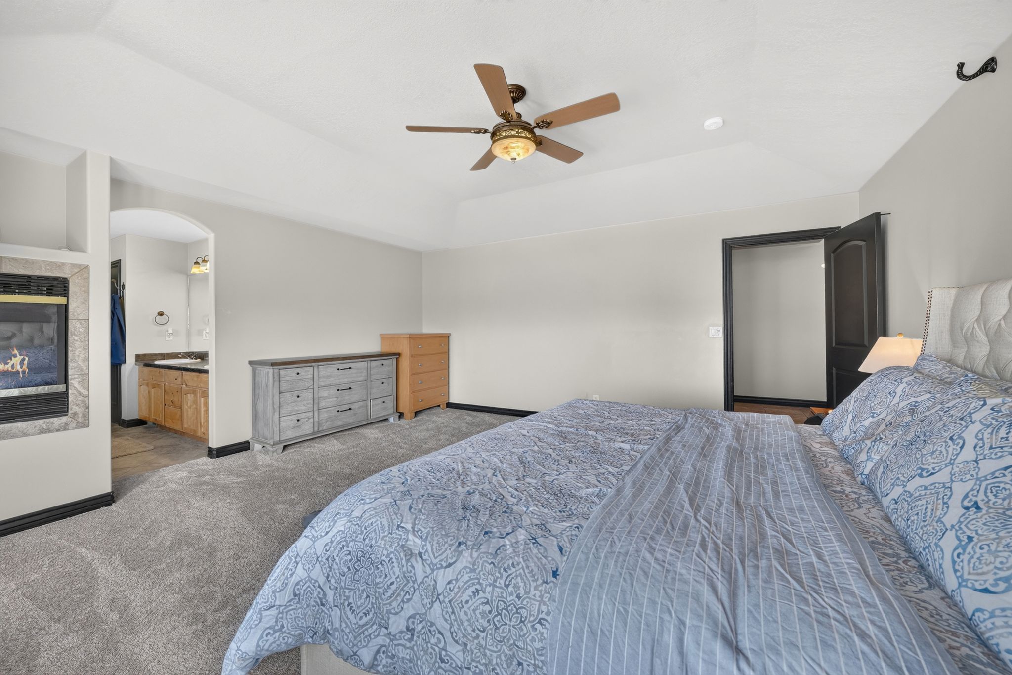Carpeted bedroom featuring a tray ceiling with a ceiling fan, a gas fireplace with a tiled surround, and an arched entry to a bathroom with a wood vanity and countertop