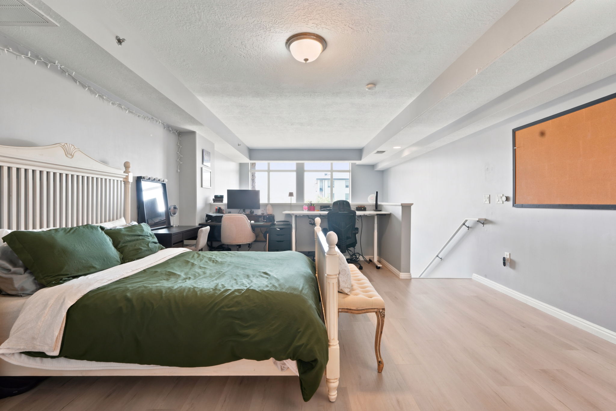 Open-concept room featuring light wood-finish flooring, a neutral wall palette, an integrated desk area, a built-in cork board, and natural light from exterior windows