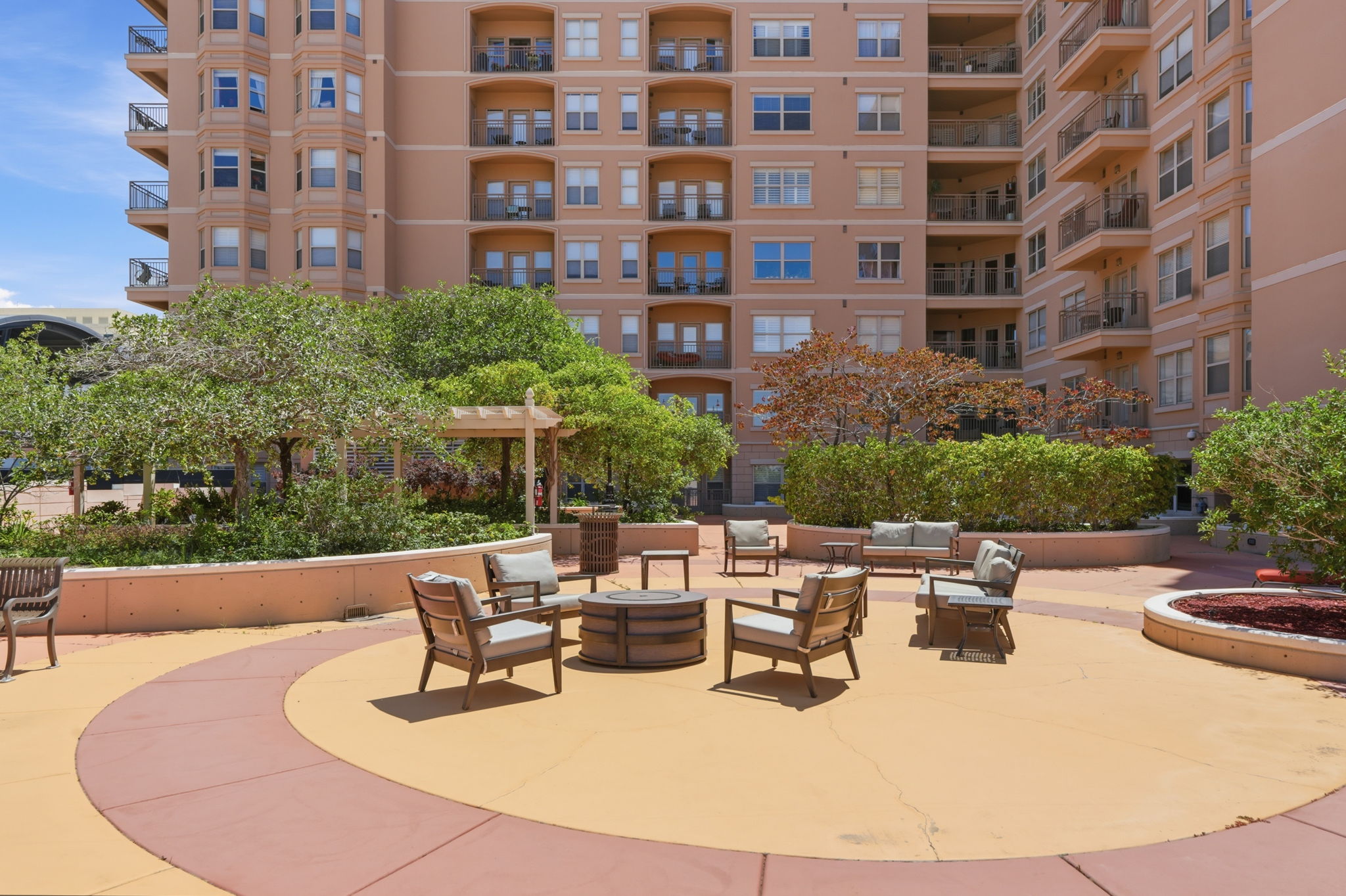 Expansive outdoor courtyard featuring patterned hardscaping, a central fire pit, established landscaping, a pergola, and seating arrangements