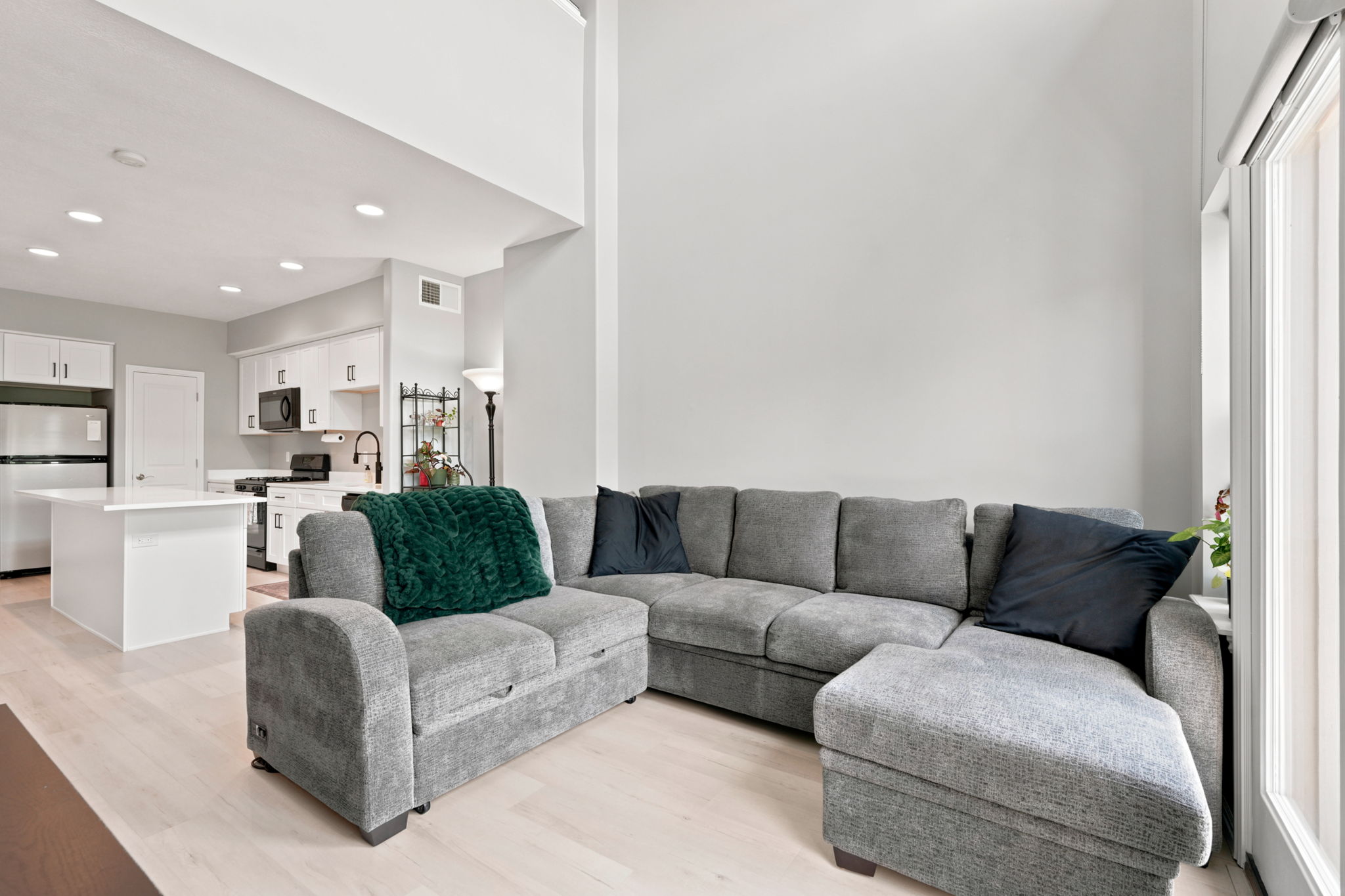 Open-concept living space featuring high ceilings, light wood-finish flooring, recessed lighting, and a kitchen with white cabinetry and stainless steel appliances