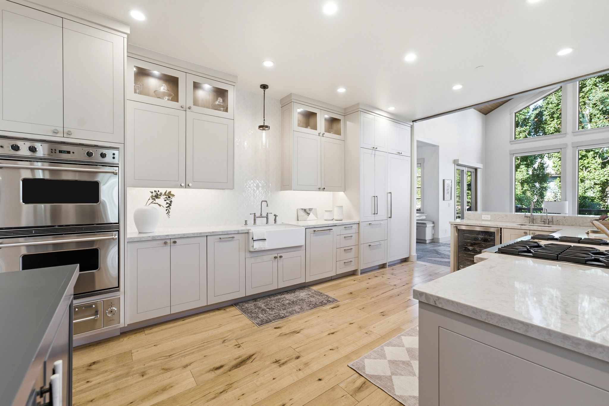 Gourmet kitchen featuring light wood-finish flooring, white cabinetry, stainless steel double ovens, a farmhouse sink, and a large center island with a cooktop and stone countertop