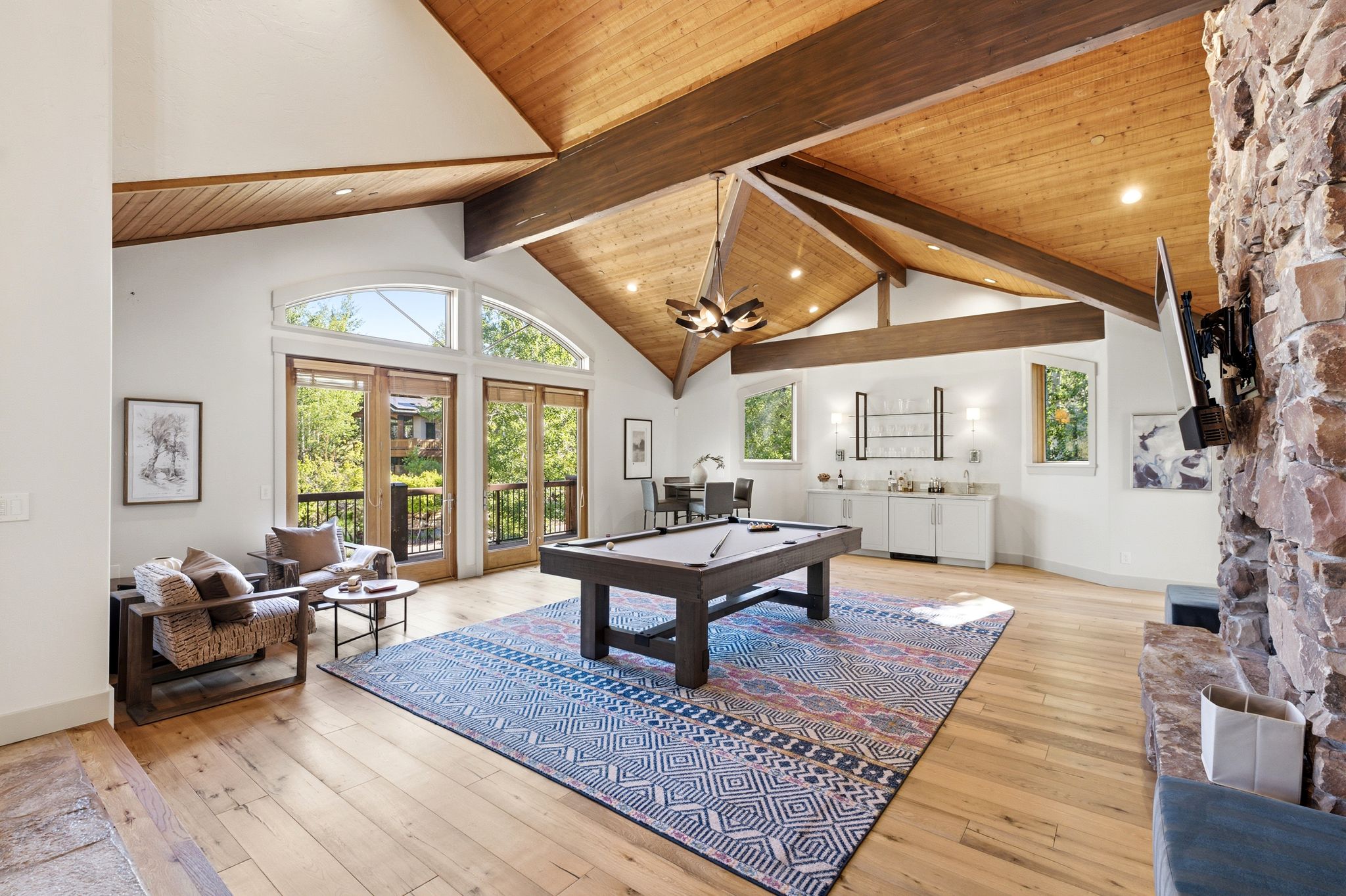 Vaulted wood-paneled ceilings with exposed beams, hardwood flooring, stone fireplace, built-in wet bar, and arched transom windows