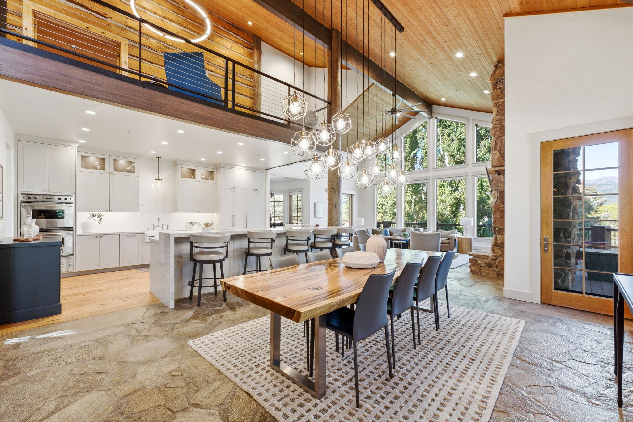 Open-concept living space featuring a modern kitchen with double ovens and an island with seating, a dining area with a linear chandelier, a vaulted wood-paneled ceiling, a stone fireplace, and large windows