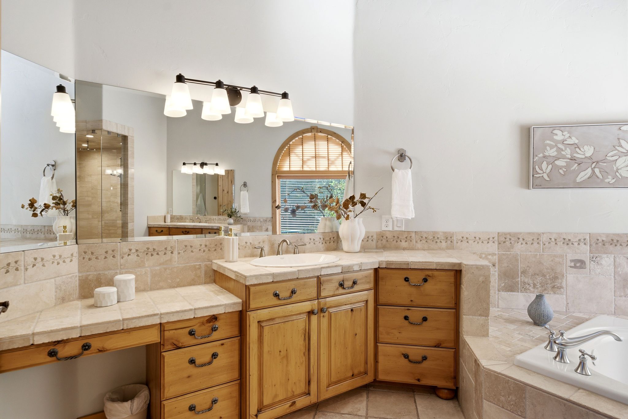 Expansive bathroom featuring natural wood cabinetry, a built-in vanity area, a tiled countertop with an integrated sink, a large wall-mounted mirror, and a soaking tub