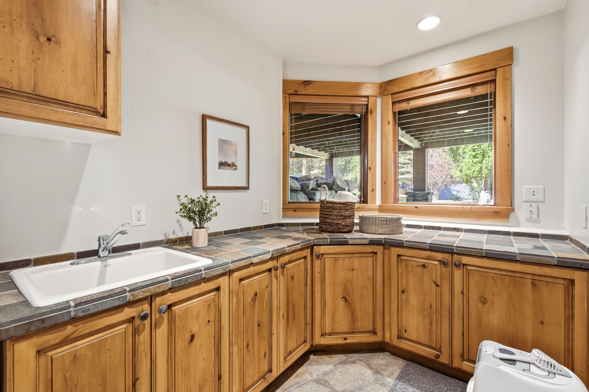 Utility room featuring knotty wood cabinetry, tile countertops, an undermount sink with chrome faucet, and two corner windows with wood trim