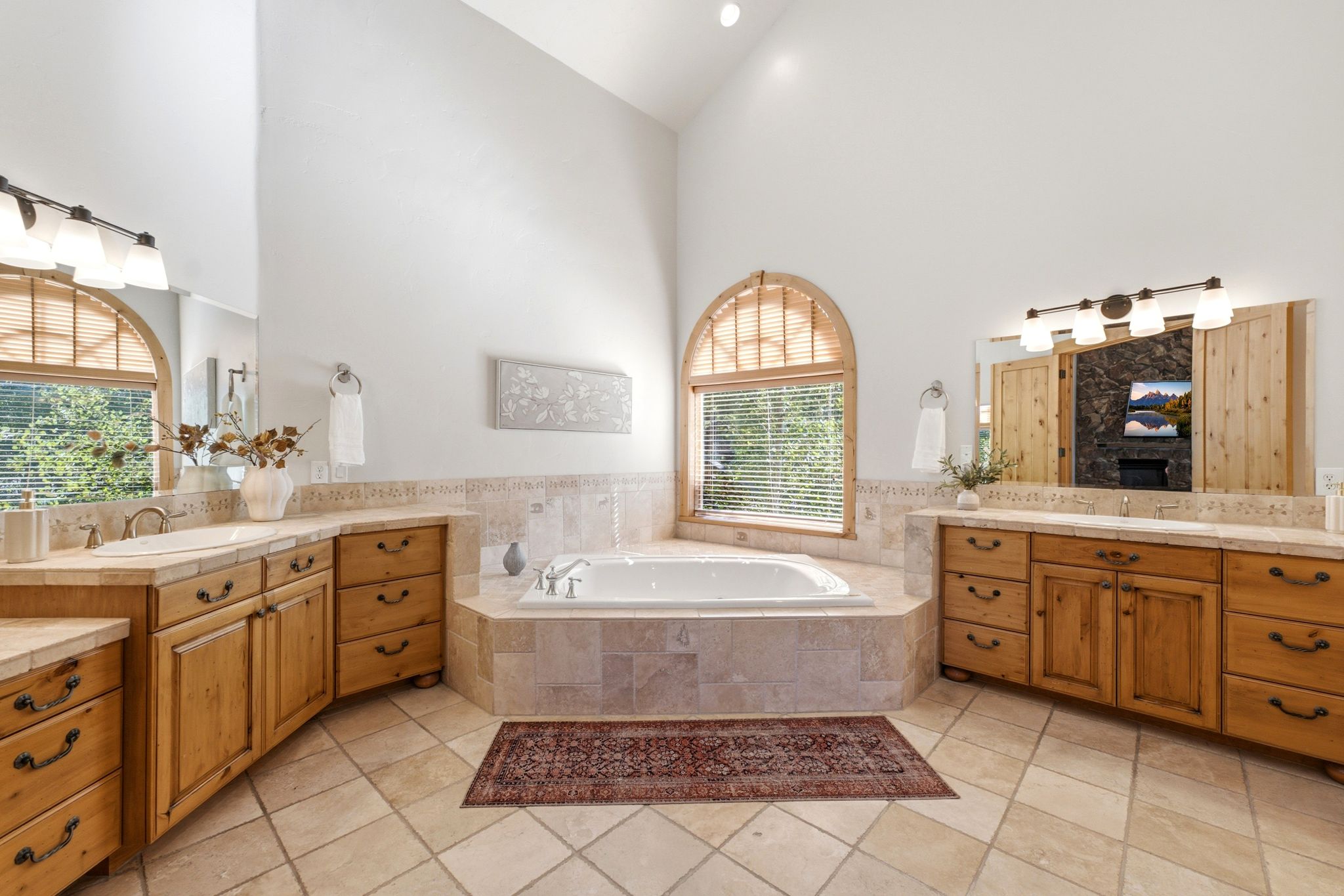 Spacious bathroom featuring a corner soaking tub, arched window with wood blinds, double vanity with wood cabinetry, tile flooring, and vaulted ceiling