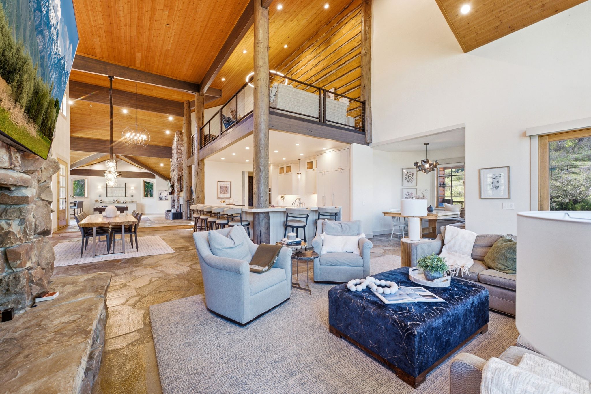 Vaulted wood-paneled ceilings with exposed beams, stone flooring, a stone fireplace, an upper-level balcony, and recessed lighting