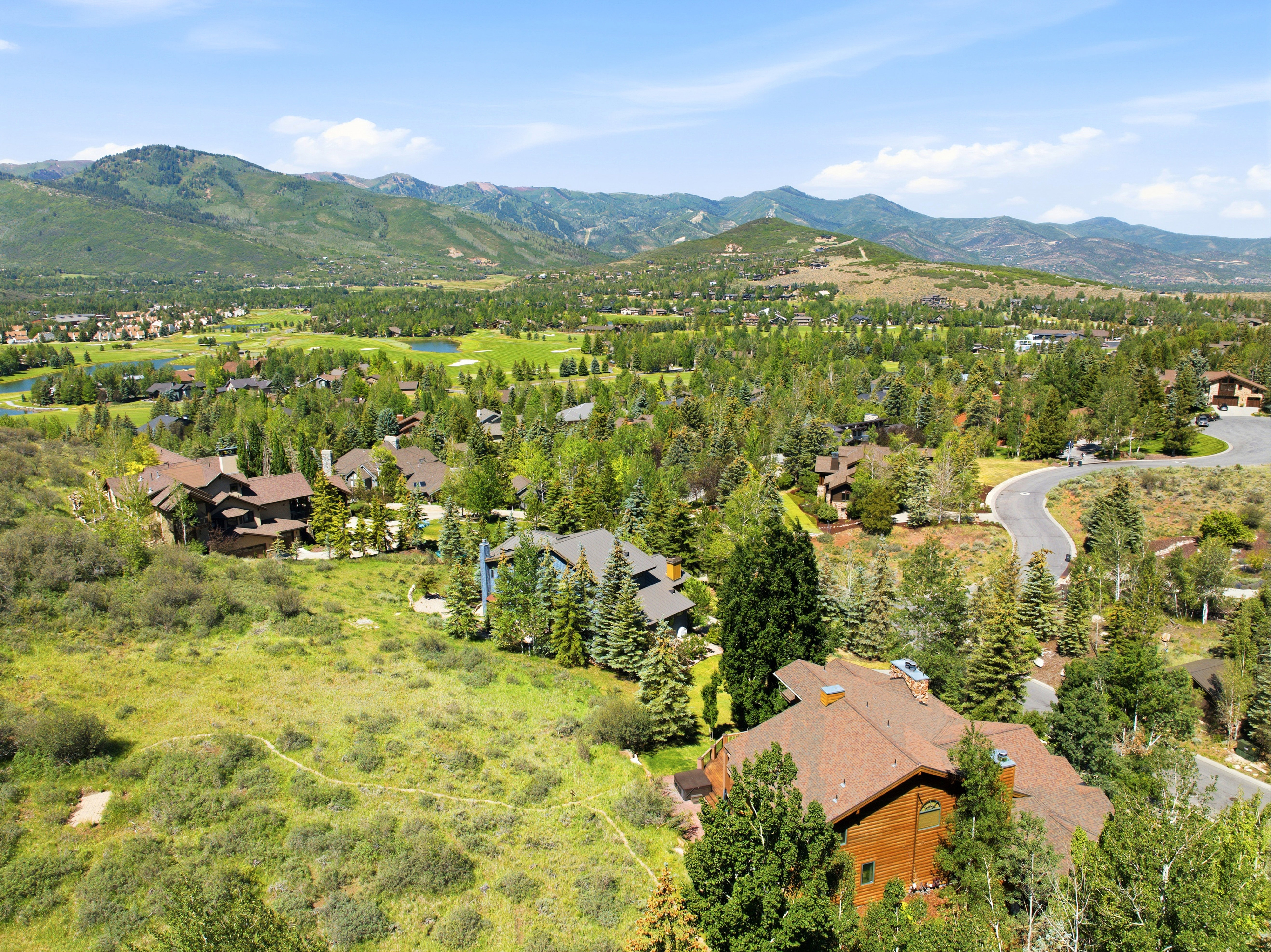 Expansive mountain landscape featuring a distant golf course, numerous mature trees, and multiple residential properties with brown-shingle roofing