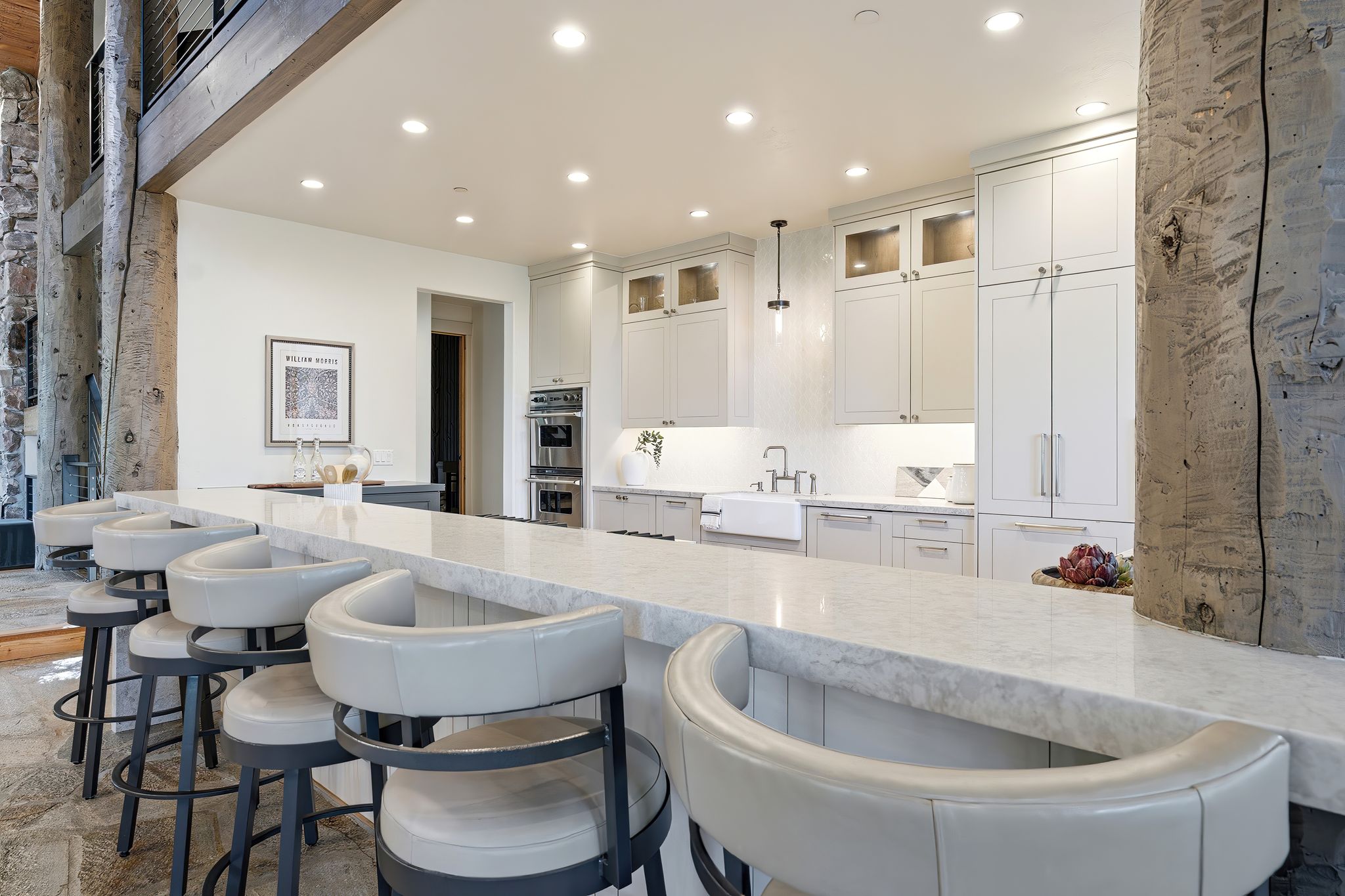 Kitchen featuring an extended island with a light-toned countertop, white cabinetry with glass upper doors, a farmhouse sink, and integrated double ovens