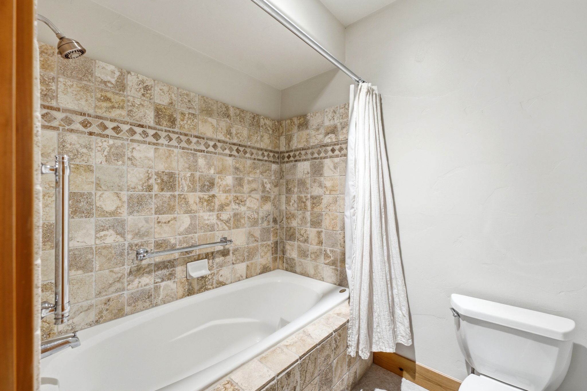 Bathtub and shower combination featuring extensive stone tile surround with decorative border, polished chrome fixtures, and a white toilet
