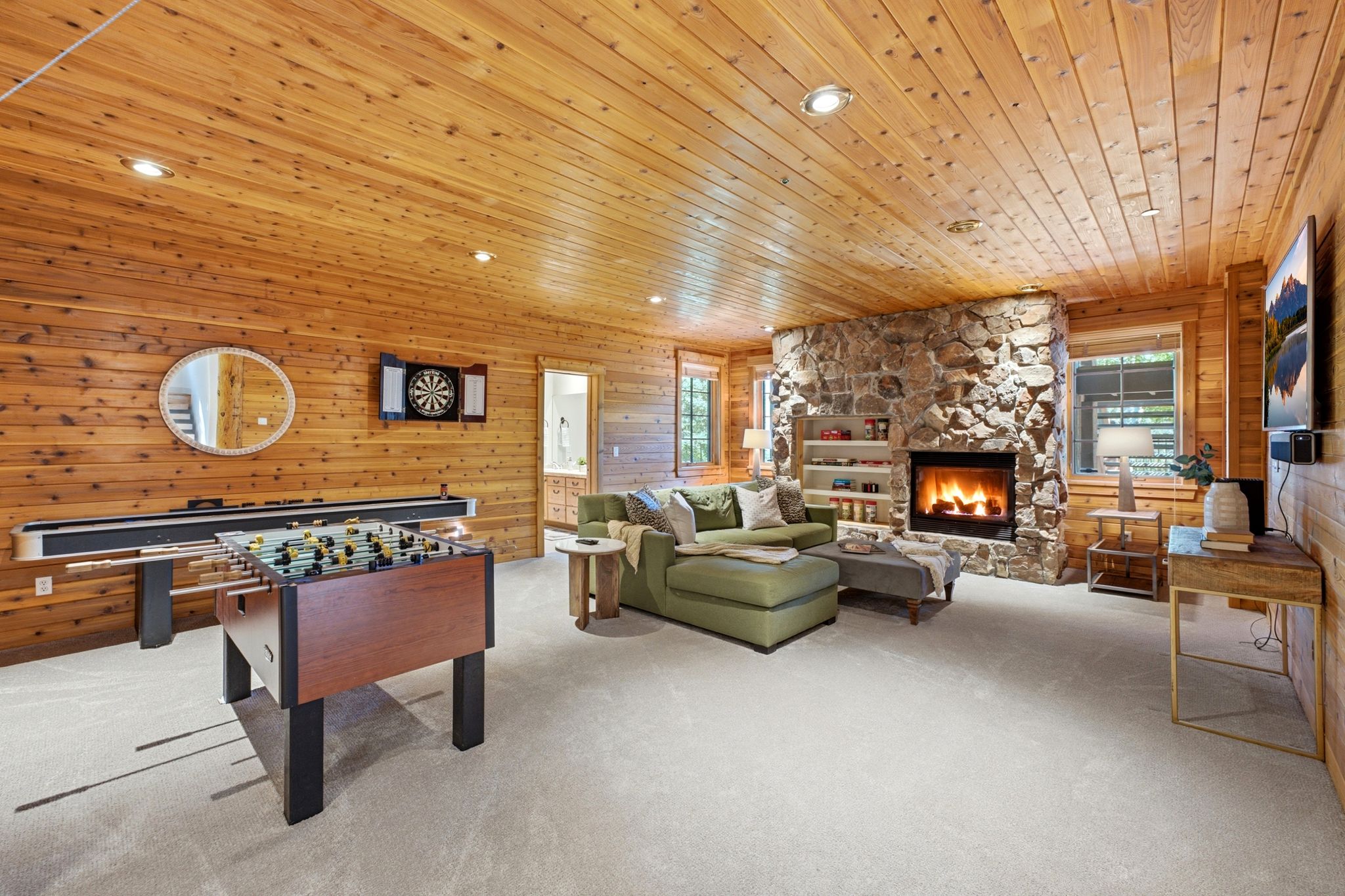 Spacious recreation area featuring wood-paneled walls and ceiling, a stone-clad fireplace with built-in shelving, recessed lighting, and light-toned carpeting