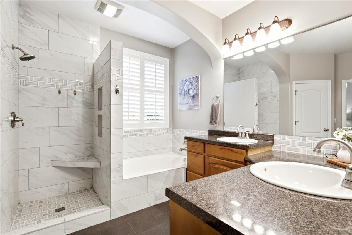 Full bathroom featuring a walk-in shower with a built-in bench and tiled niche, a separate bathtub with plantation shutters, a dual vanity with undermount sinks and dark stone-finish countertops, and a large wall-mounted mirror