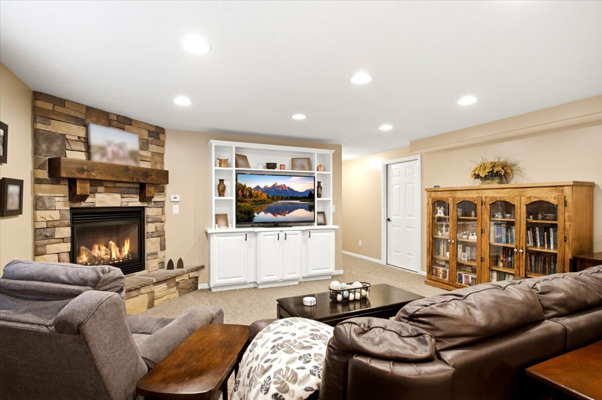 Stacked stone fireplace with a substantial wood mantel, built-in media center with shelving, recessed lighting, neutral carpeting, and a white paneled door