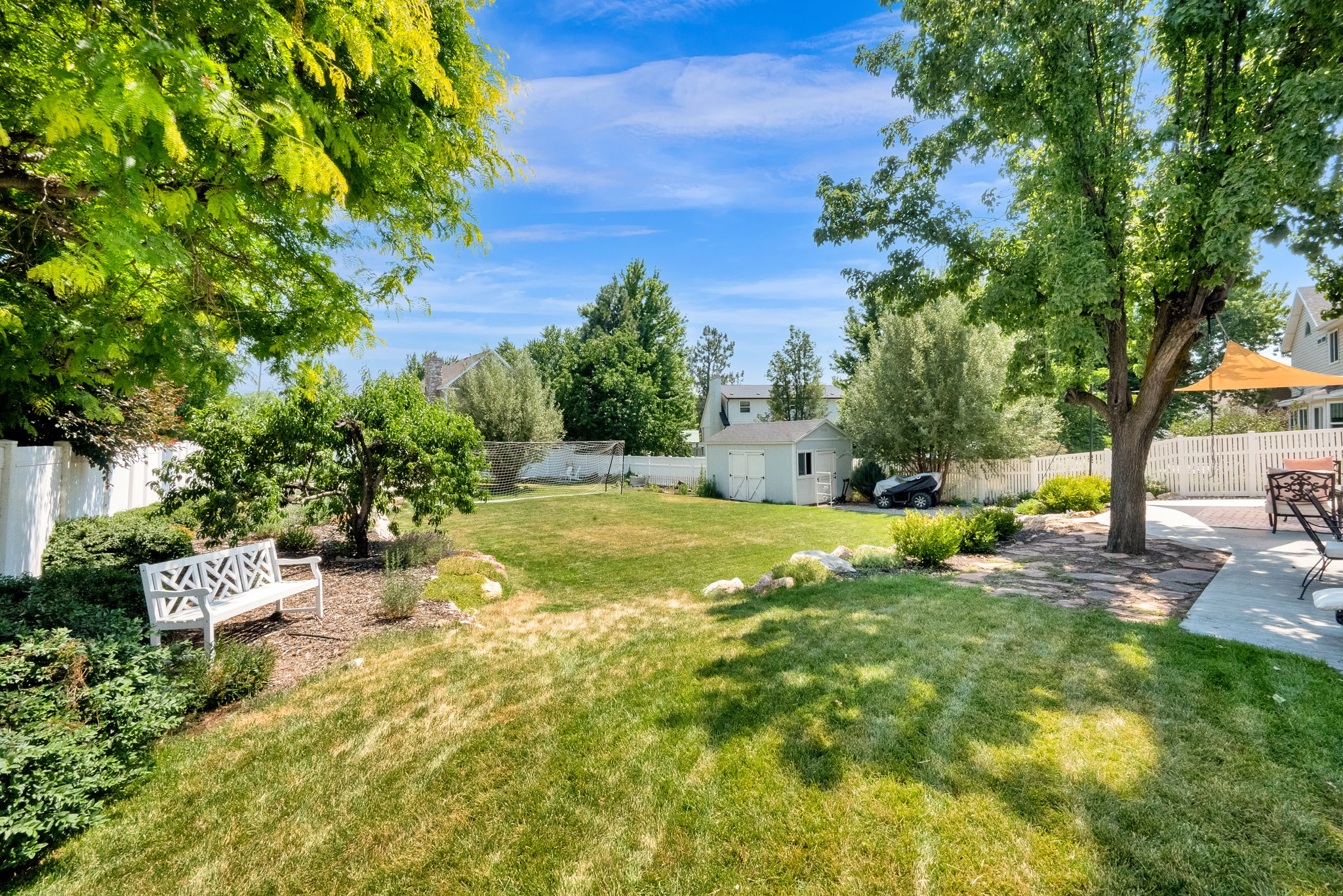 Expansive backyard featuring a lush lawn, mature trees, white picket fencing, a concrete patio, and a storage shed