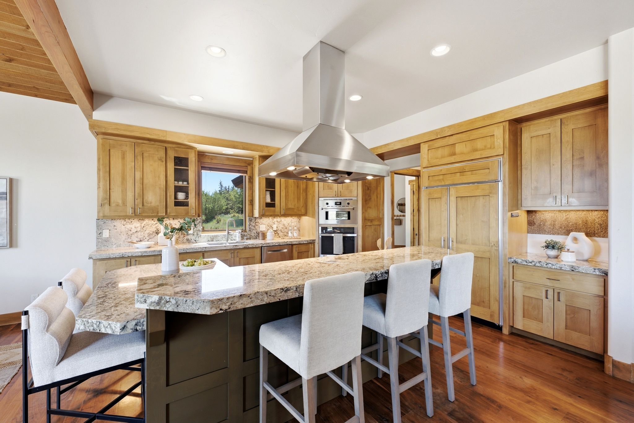 Spacious kitchen featuring a large island with a stone countertop, built-in stainless steel appliances, custom wood cabinetry, wood-finish flooring, and exposed wood ceiling beams