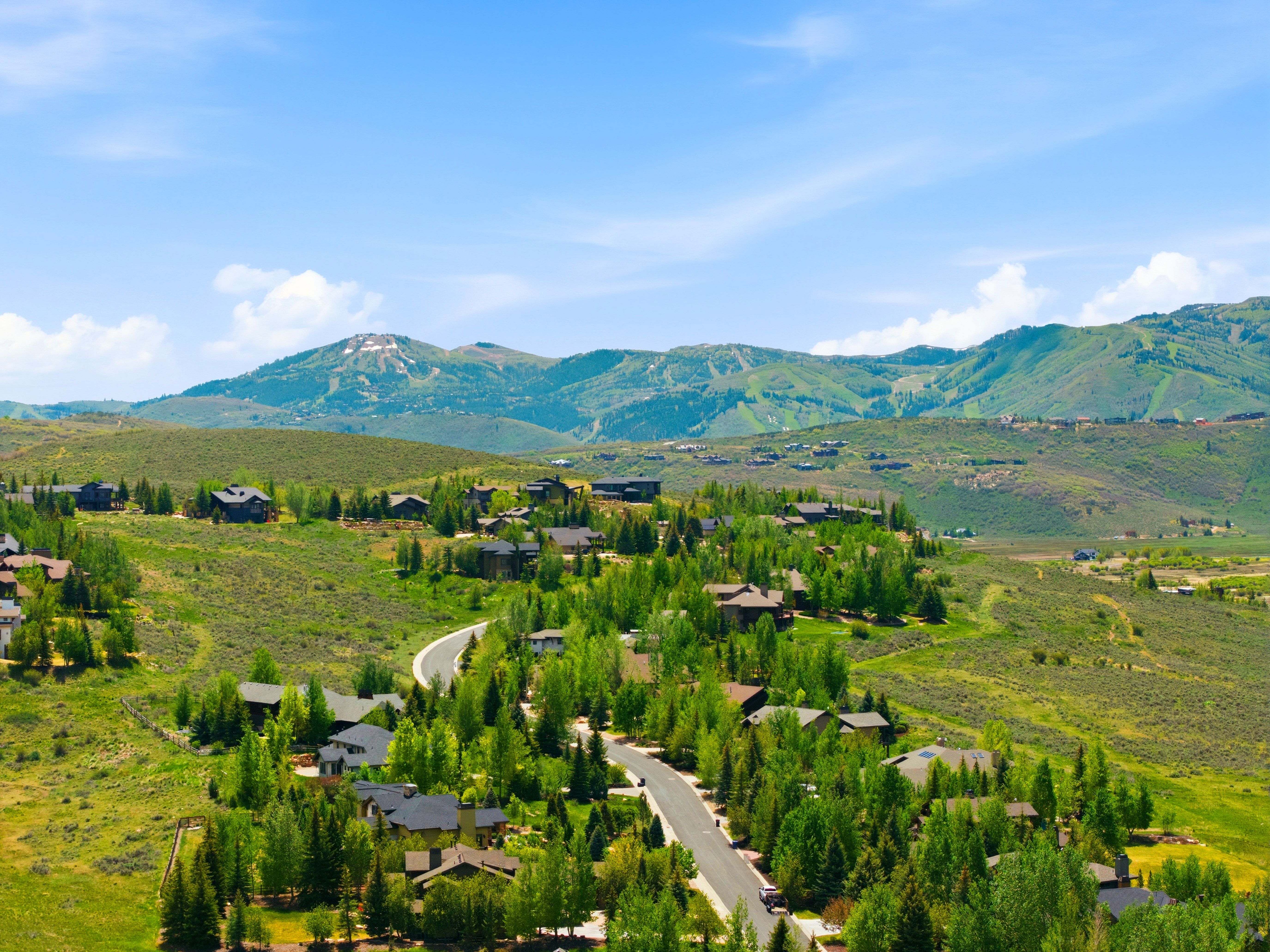 Expansive mountain landscape featuring varied terrain with distant peaks, a clear blue sky, numerous residential properties, and paved roadways