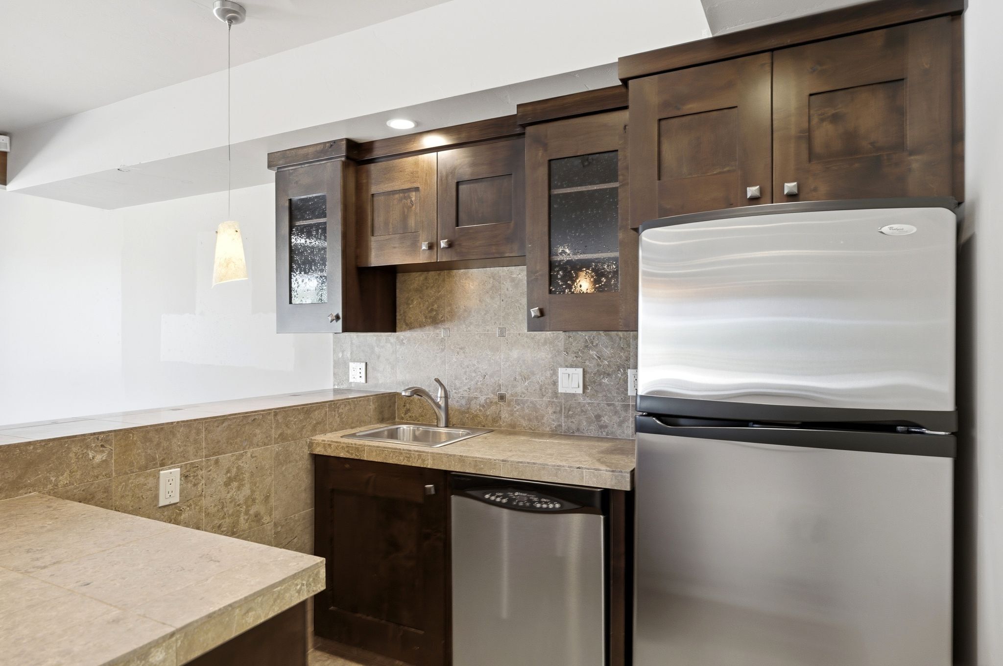 Compact kitchen area featuring wood cabinetry, stainless steel appliances, a bar sink, stone countertops, and a coordinating stone backsplash