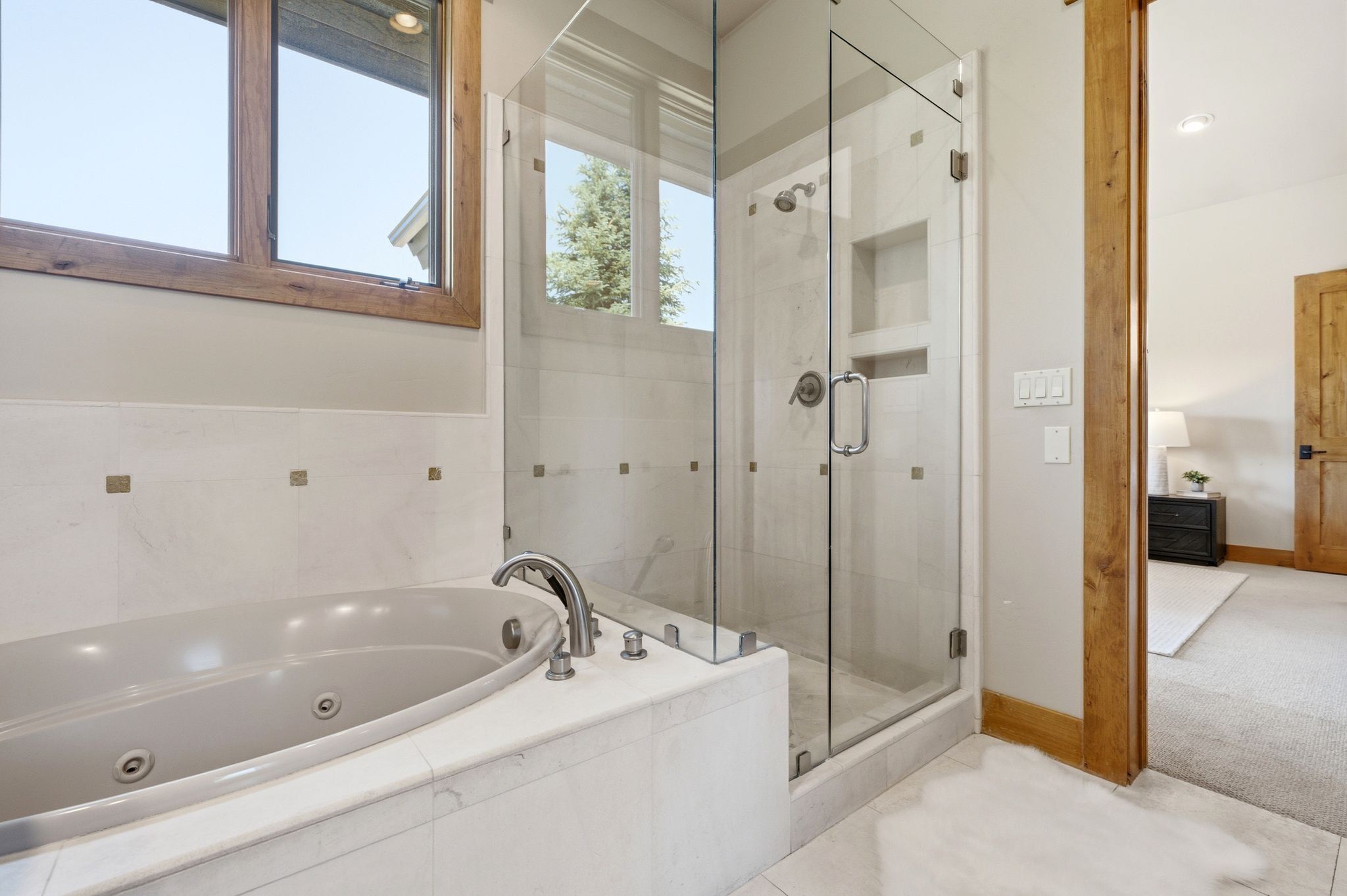 Bathroom featuring a built-in jetted tub with deck-mounted faucet, a glass-enclosed shower with tile surround and recessed shelving, and light wood-frame windows