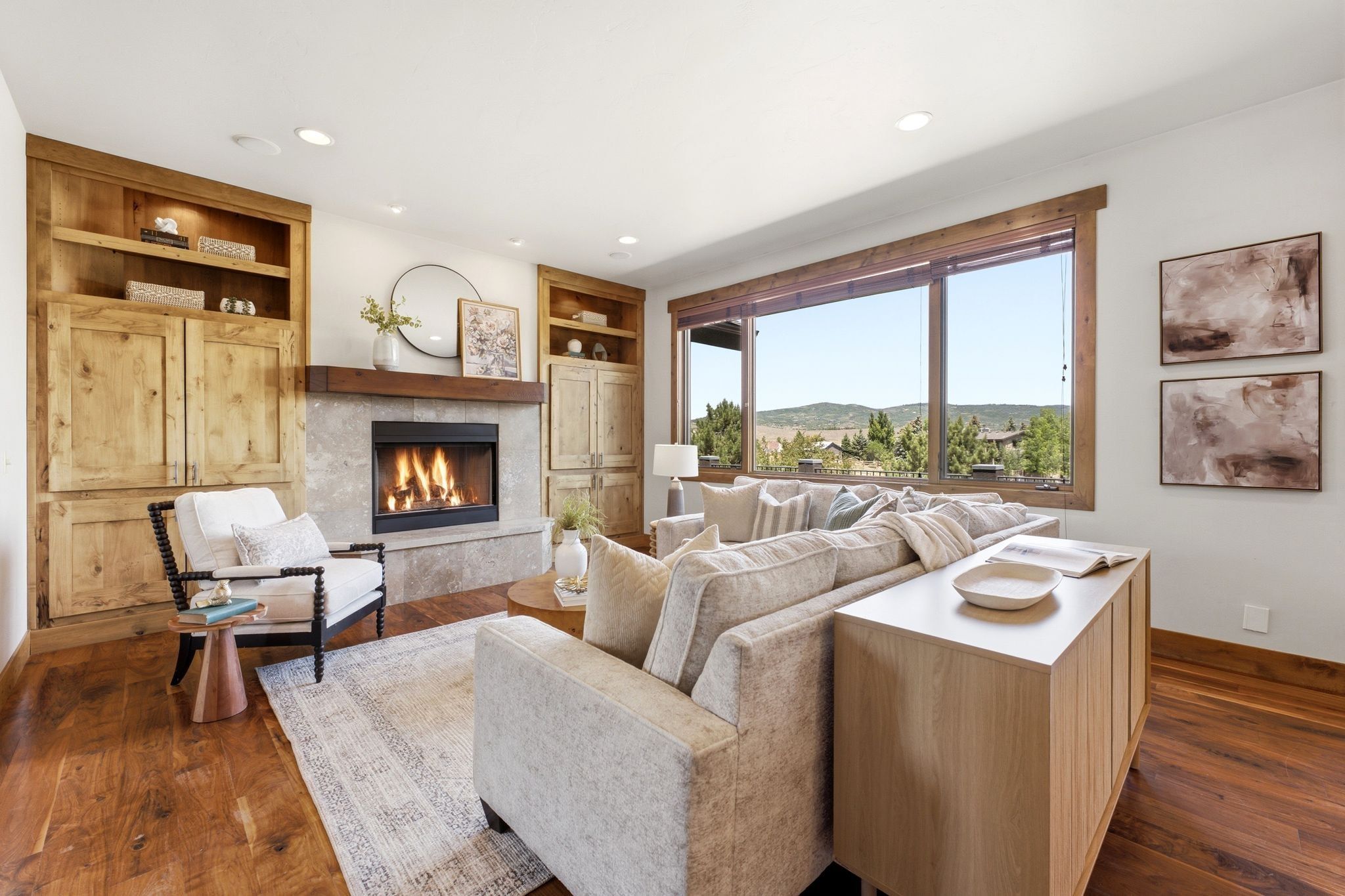 Living area featuring a stone fireplace with a wood mantle, built-in wooden cabinetry, expansive window framing distant tree-covered hills, recessed lighting, and wood-finish flooring