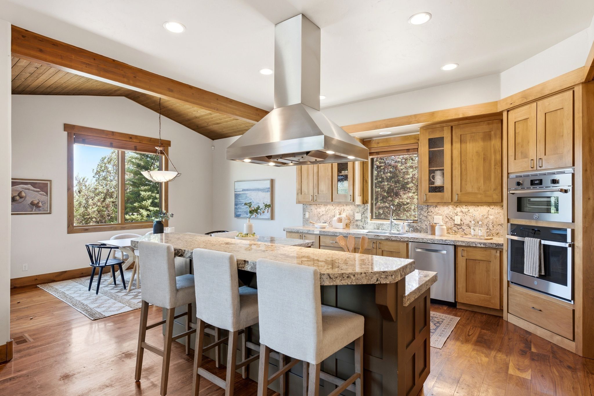 Spacious kitchen featuring a large granite-topped island with seating, stainless steel appliances, wood-finish flooring, exposed wood beams, and ample cabinetry