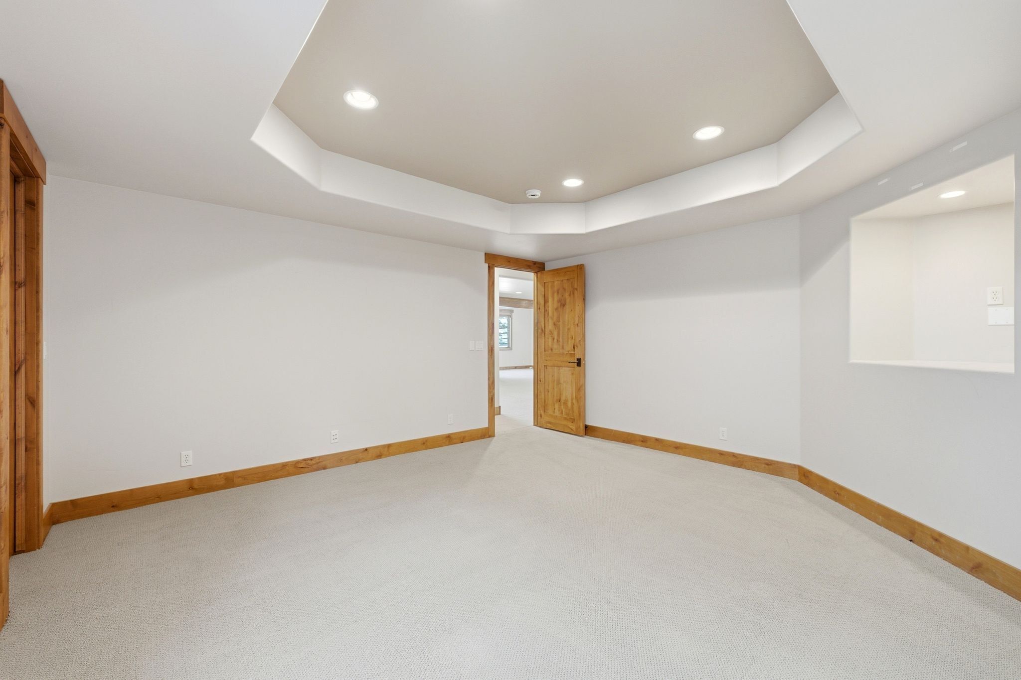 Spacious room featuring a tray ceiling with recessed lighting, light-colored carpet, natural wood trim, and a built-in wall niche