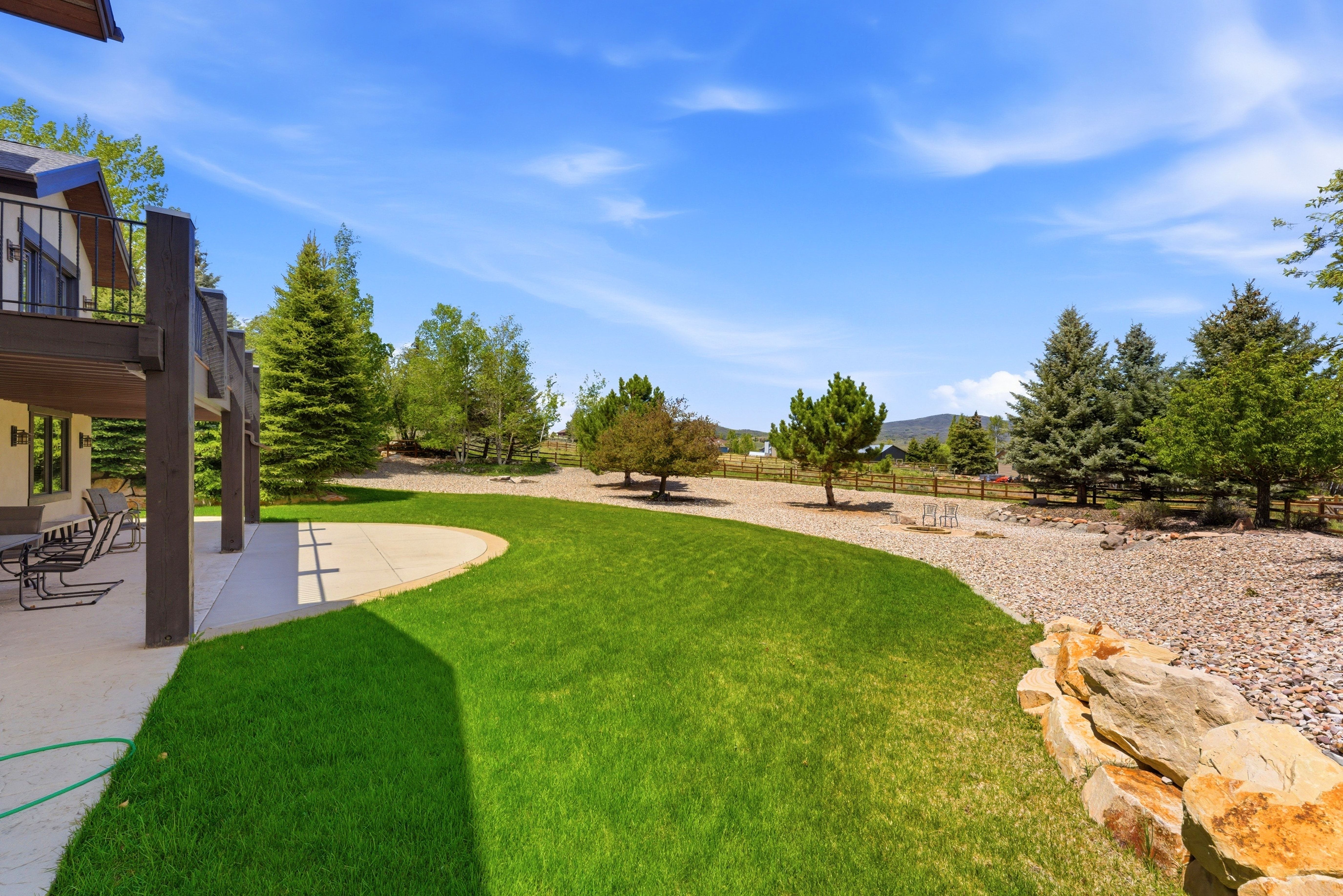 Expansive rear yard featuring a curved concrete patio, lush green lawn, natural rock landscaping, mature evergreen trees, and a distant mountain backdrop