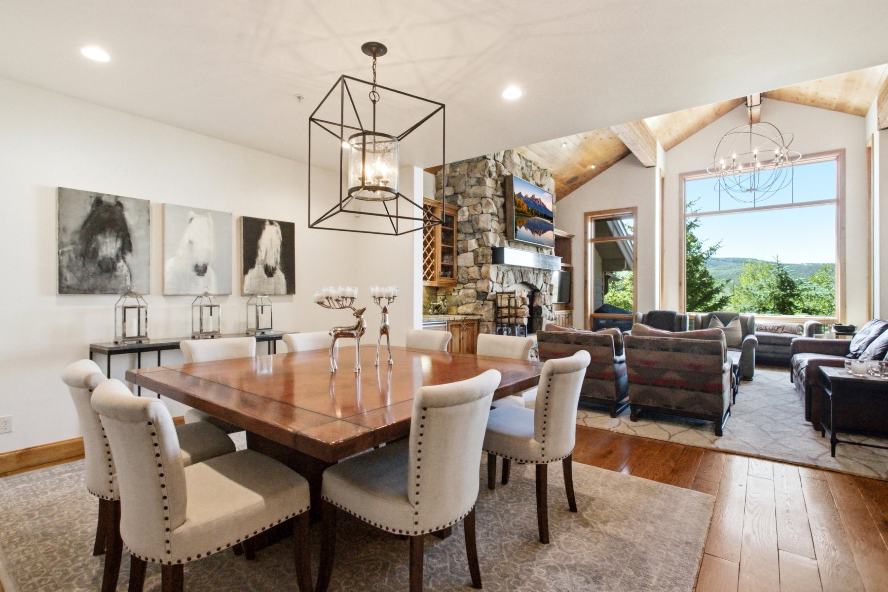 Open-concept living space with vaulted ceilings featuring exposed wood beams, a stone-clad fireplace, and large picture windows