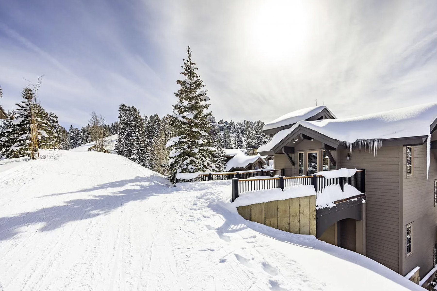 Snow-covered ski path bordering a contemporary multi-level residence with dark siding, a private balcony, and an exposed concrete retaining wall