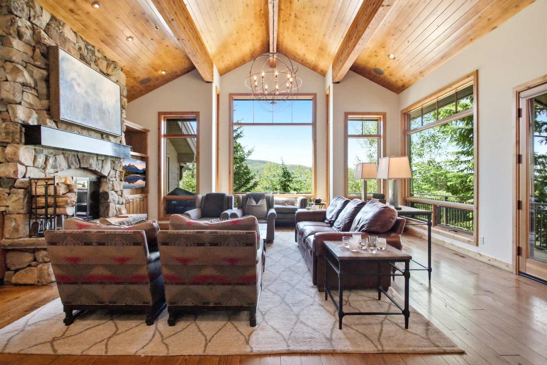 Spacious living area featuring a stone fireplace with a substantial hearth, vaulted wood-plank ceilings with exposed beams, and expansive windows