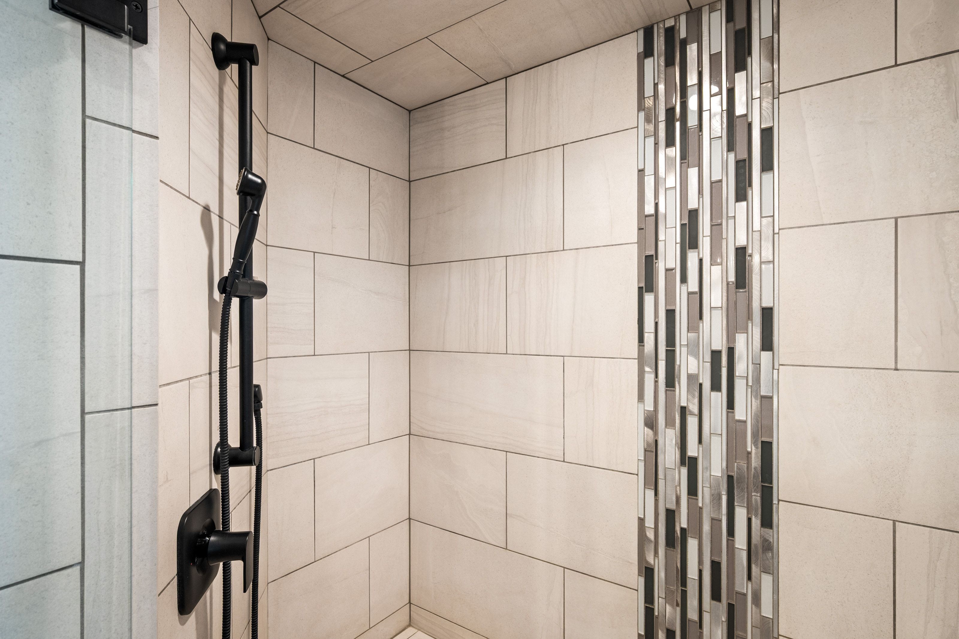 Shower stall featuring large format rectangular wall tiles, a vertical accent strip of mosaic tile, and a matte black shower fixture with a handheld spray