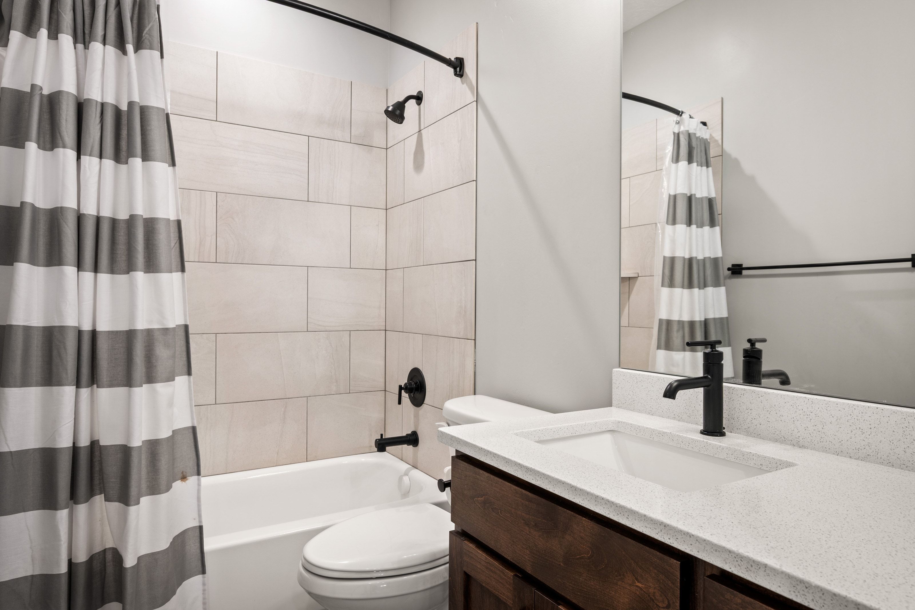 Bathroom featuring a shower/tub combination with light-toned rectangular tile surround, matte black fixtures, a dark wood-finish vanity with an integrated sink, and a speckled light countertop