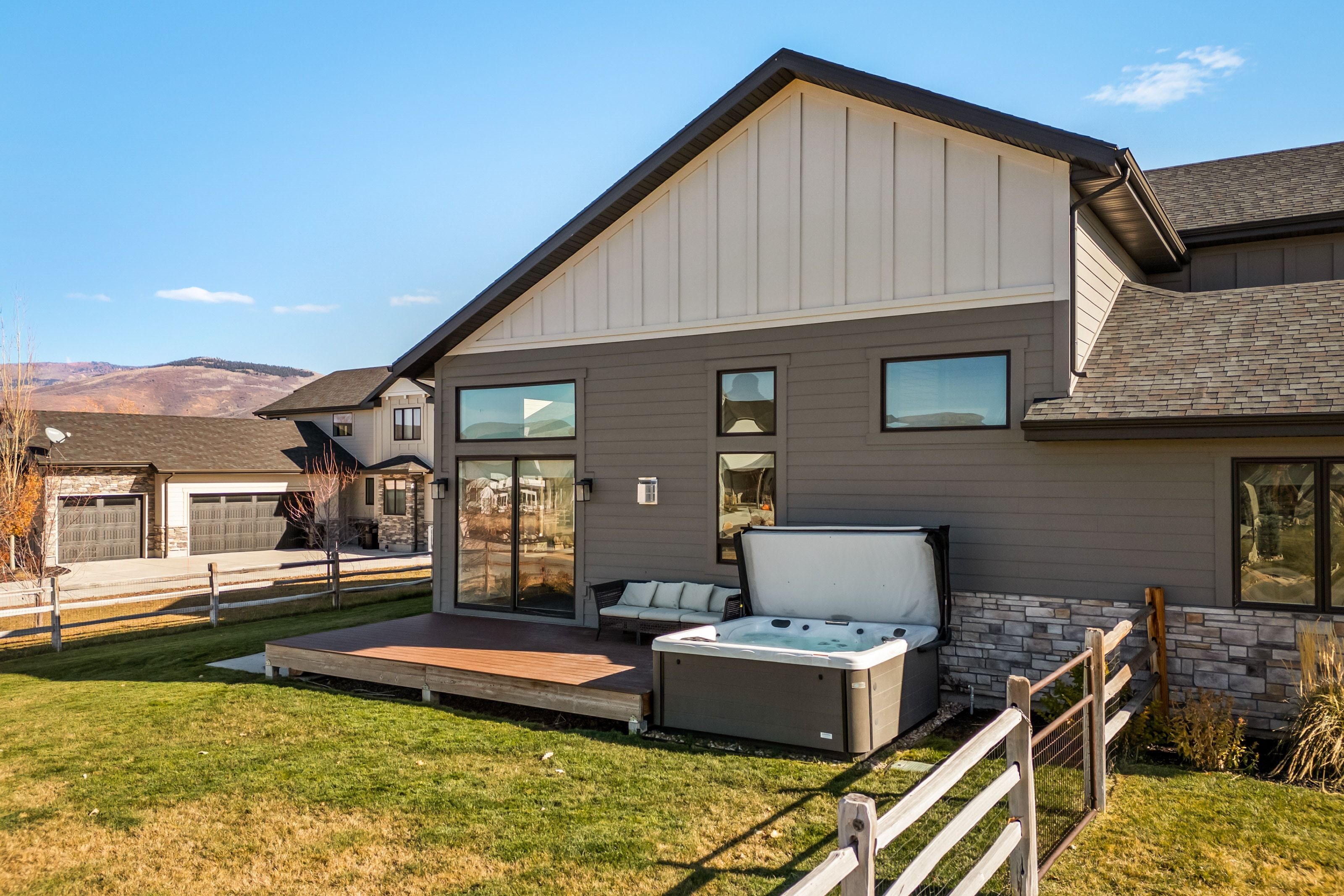 Rear exterior featuring a hot tub on a wood-finish deck, vertical and horizontal siding, stone veneer accents, and large windows